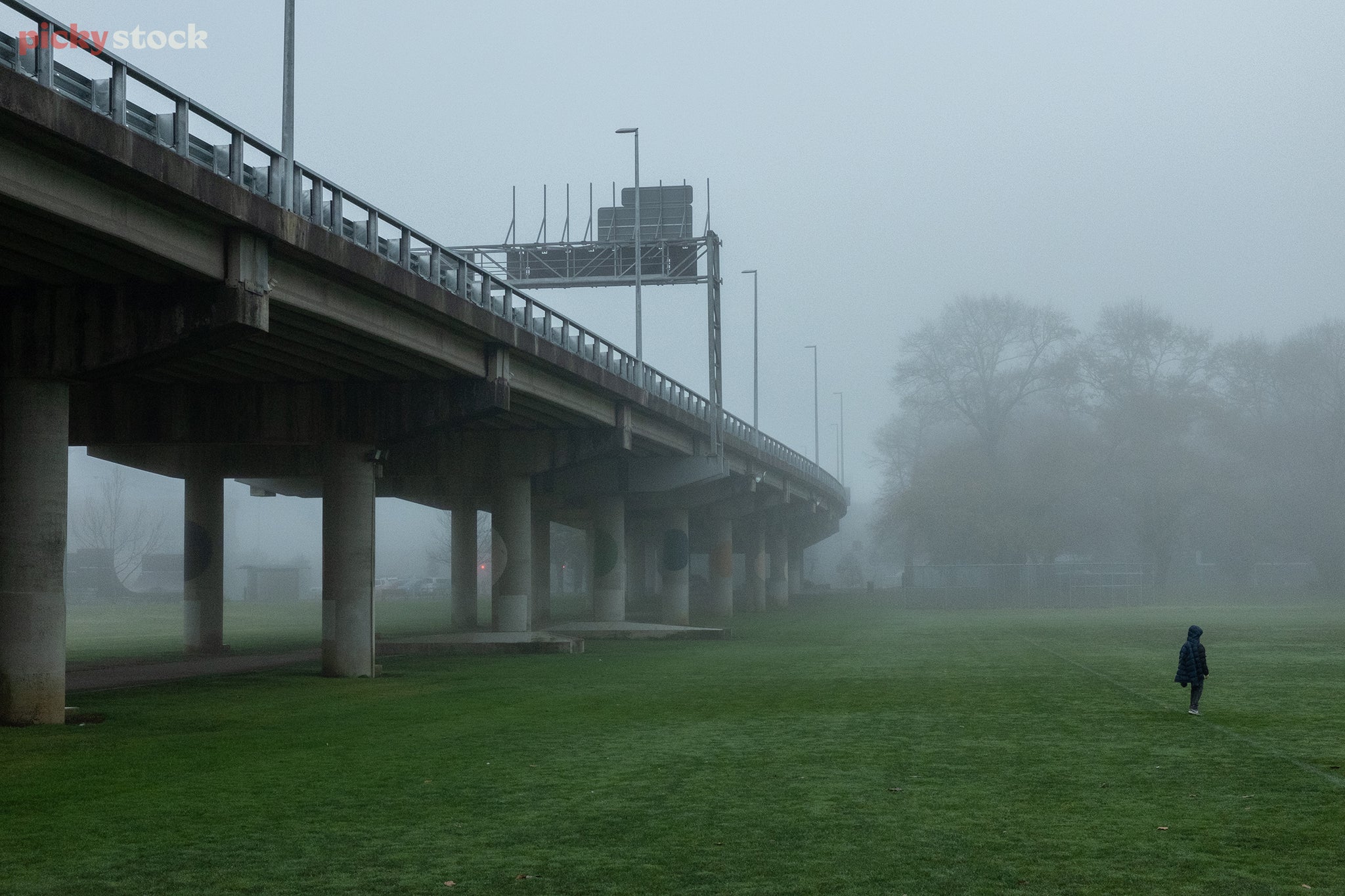 Landscape is a concrete overpass stretching high above green sports fields. A lone watcher in a dark blue puffer jackets stares into the distance as grey fog lurks around.