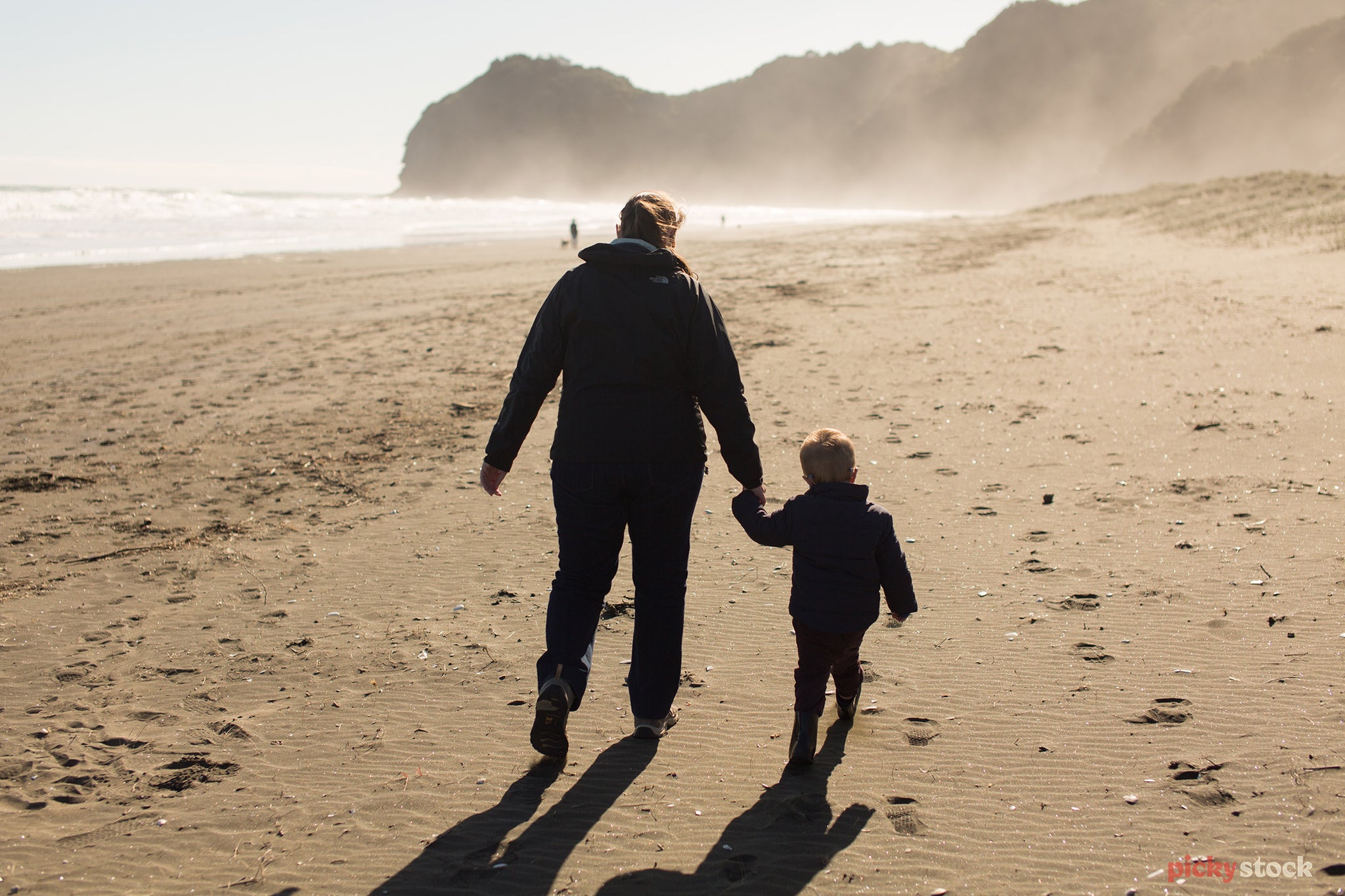 Parent and child hold hands walking along beach facing back to camera. Middle of the day. black sand beach with a mild sea spray visible. 
