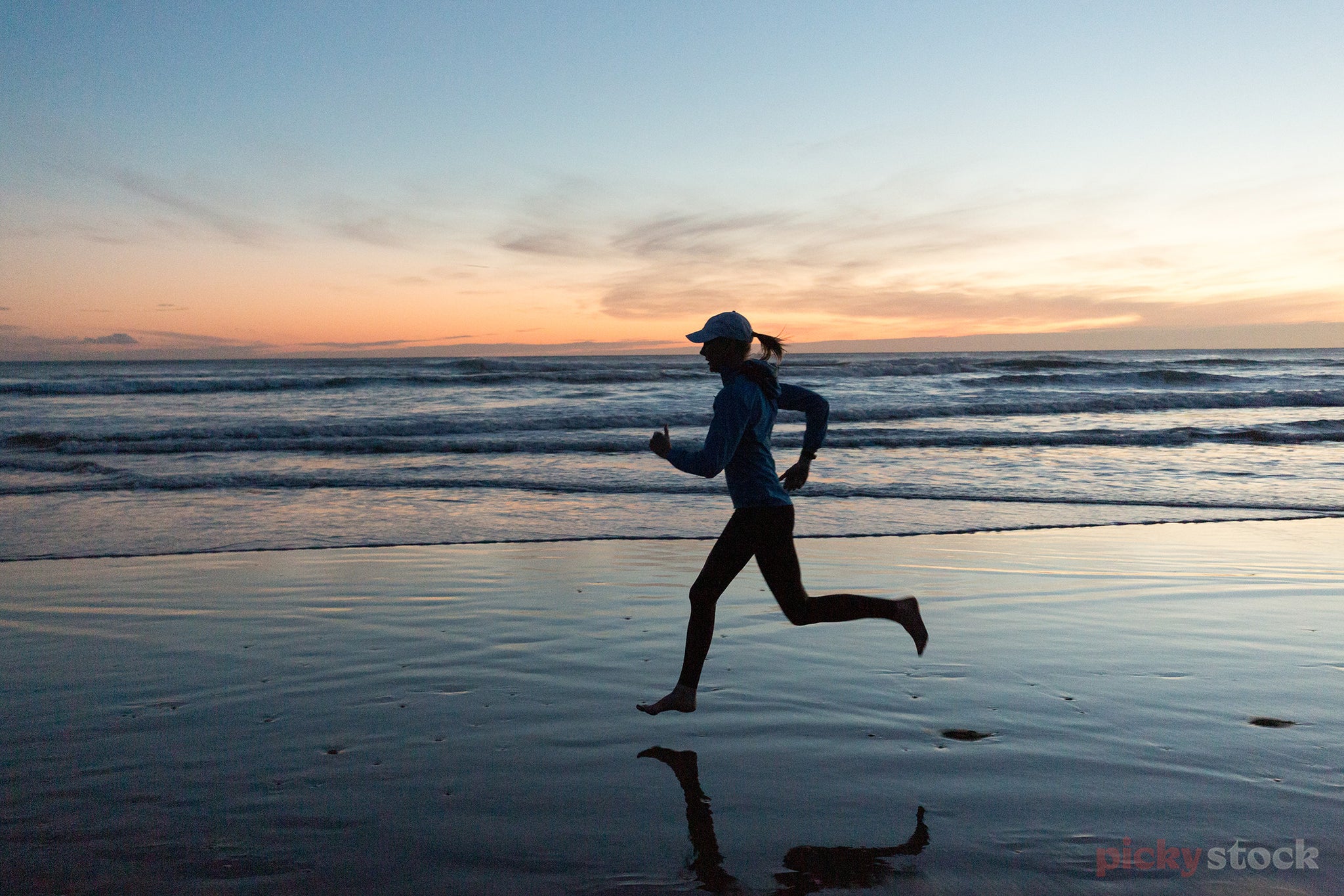 Lady runs along beach at dawn / dusk.  Sky is a dark blue with a bright orange along horizon line. Lady is centre of frame as a black outline. 