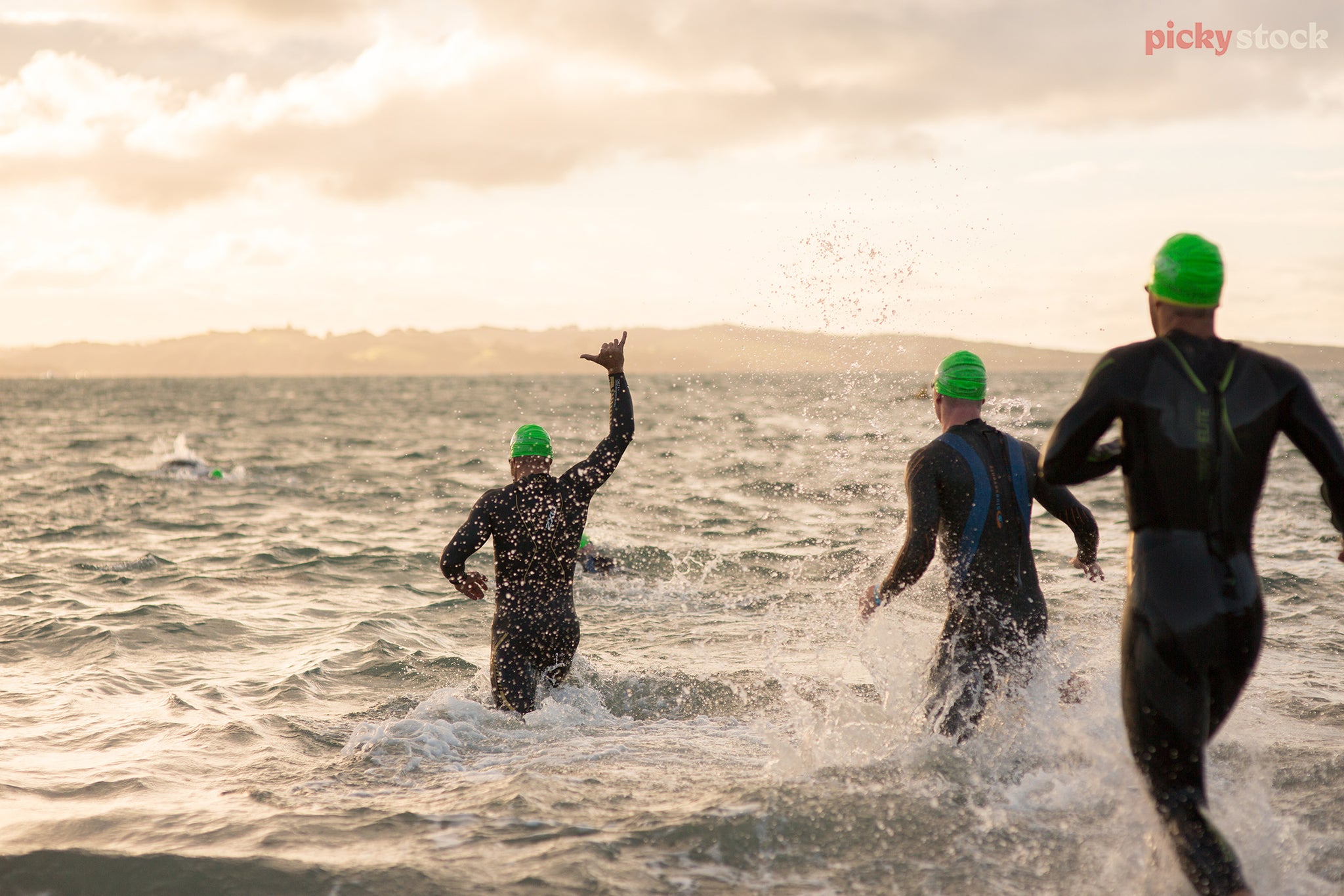 Group of ocean swimmers run into water, ready to swim. Wearing wet suits and bright green swimming caps. Sky is golden. 
