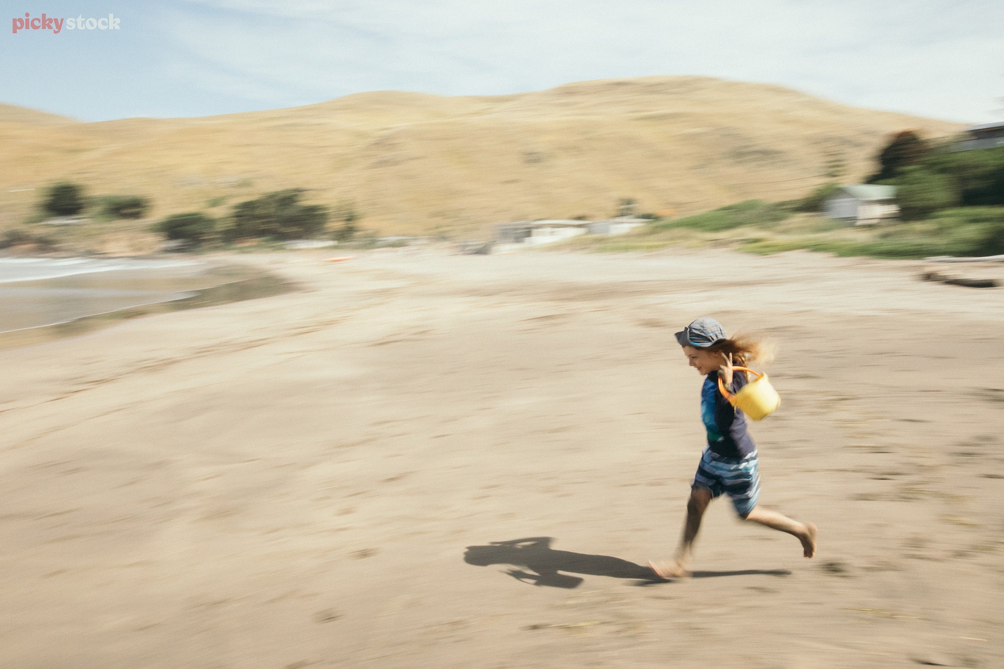 Child speedily runs along beach to water carrying a bright yellow bucket. Image has a motion blurr. Background is a soft beach day, blue summer sky. 