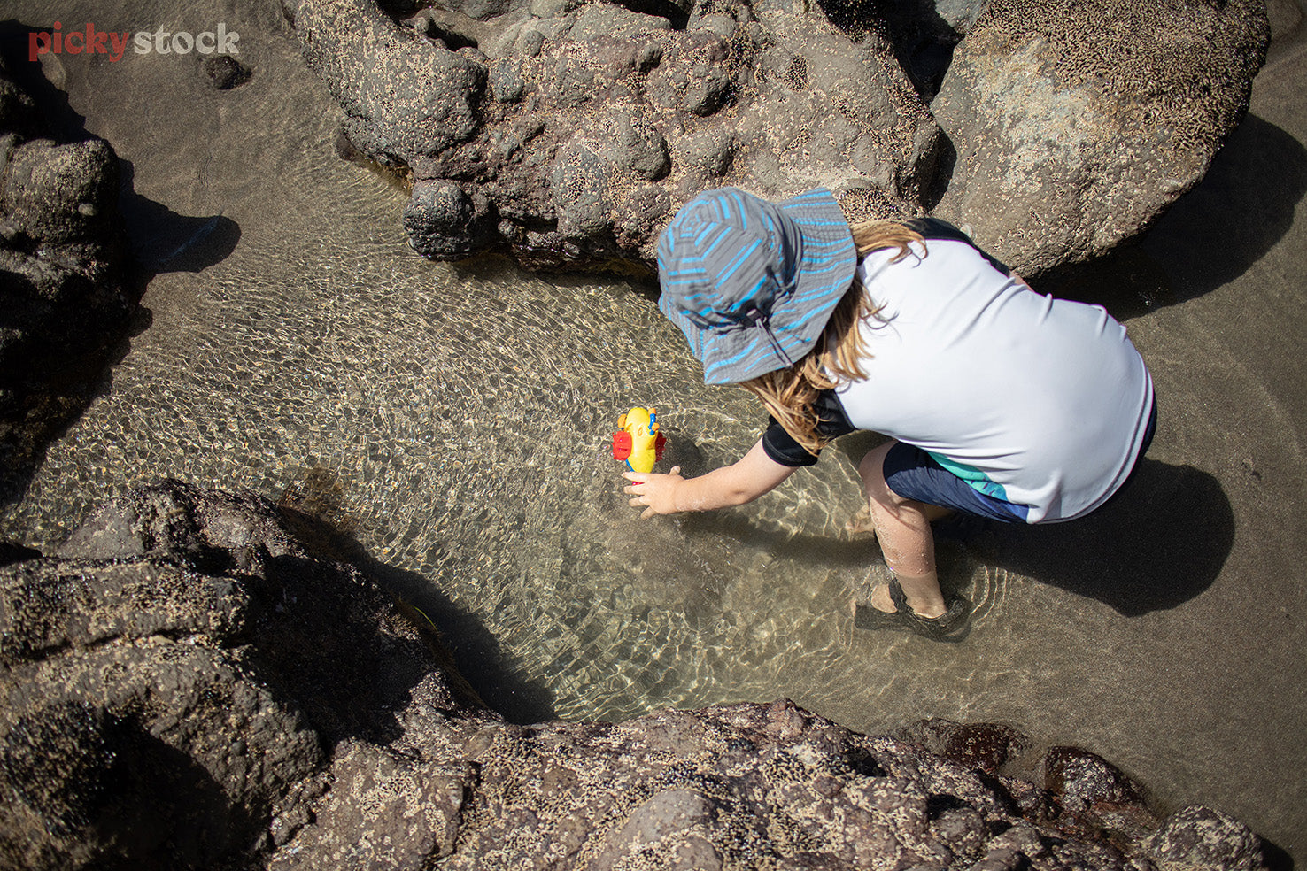 Small girl playing with toy submarine in the rock pools and shallow water. As seen from above. 