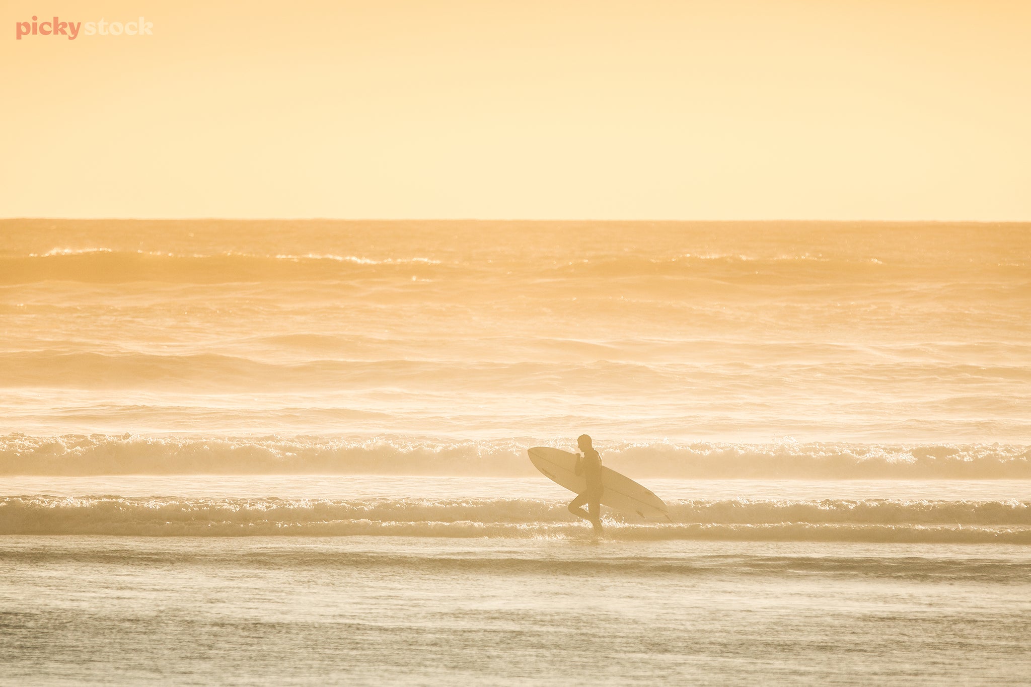 Landscape of a male surfer in a black wetsuit ruing across the beach as the tide breaks upon the shore, the hazy light of the sun blurs the image like exposed film.