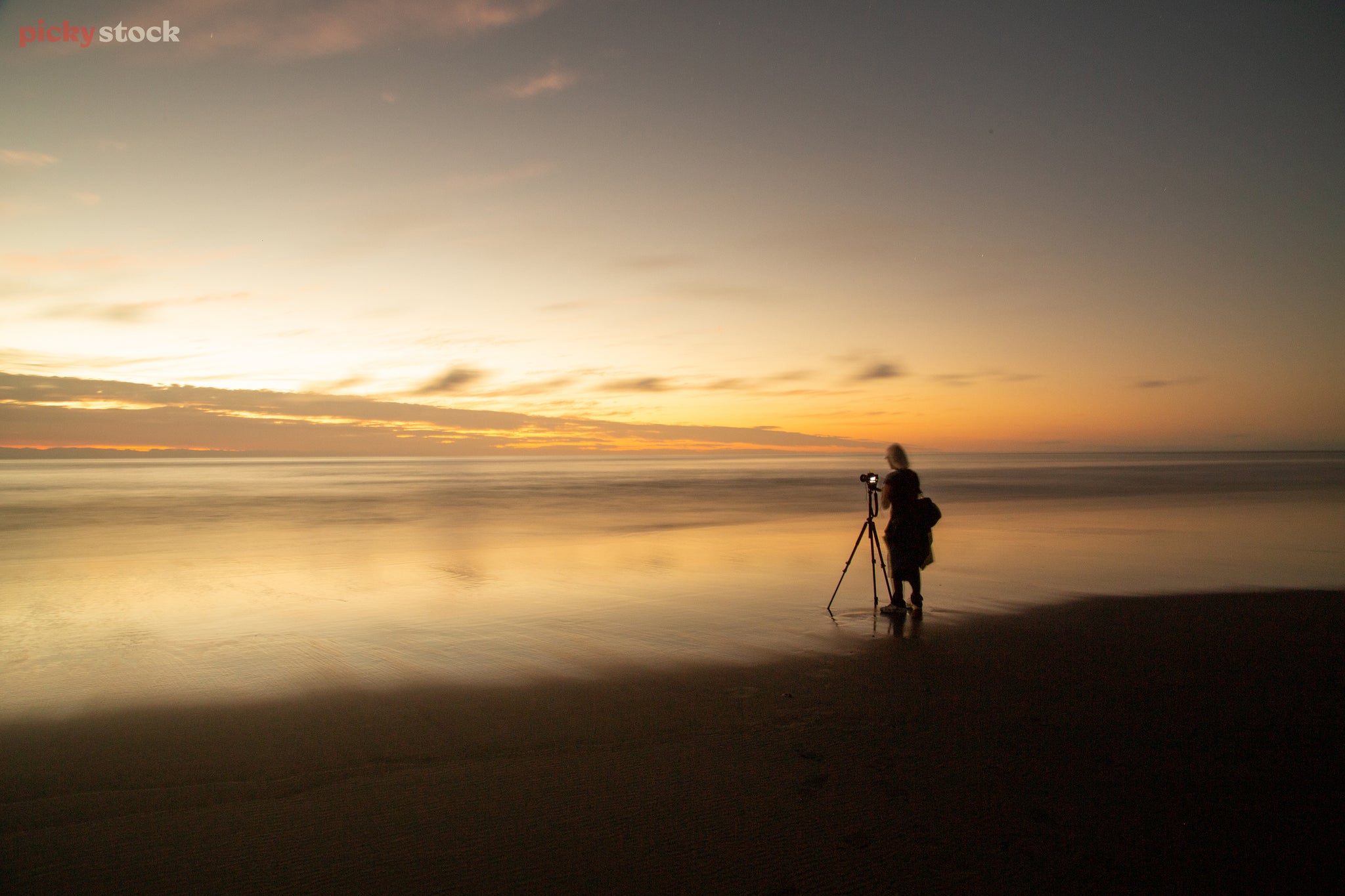 Landscape of a woman photographing the horizon from a beach, the tide has receded and the tripod buried in the sand creates a shallow bowl of water. The sun crashes like a huge gold wave against the sky.