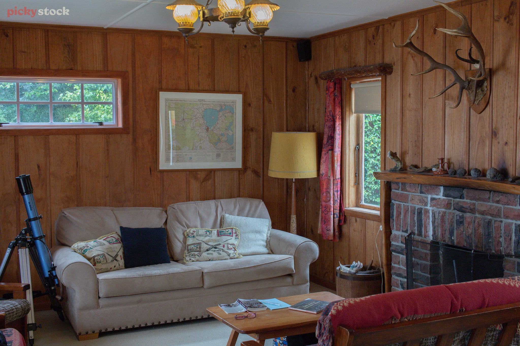 Landscape of a living room with antique furnishing and decor a set of antlers hang over a brick fireplace. A large telescope sits folded in the corner next to a framed map.