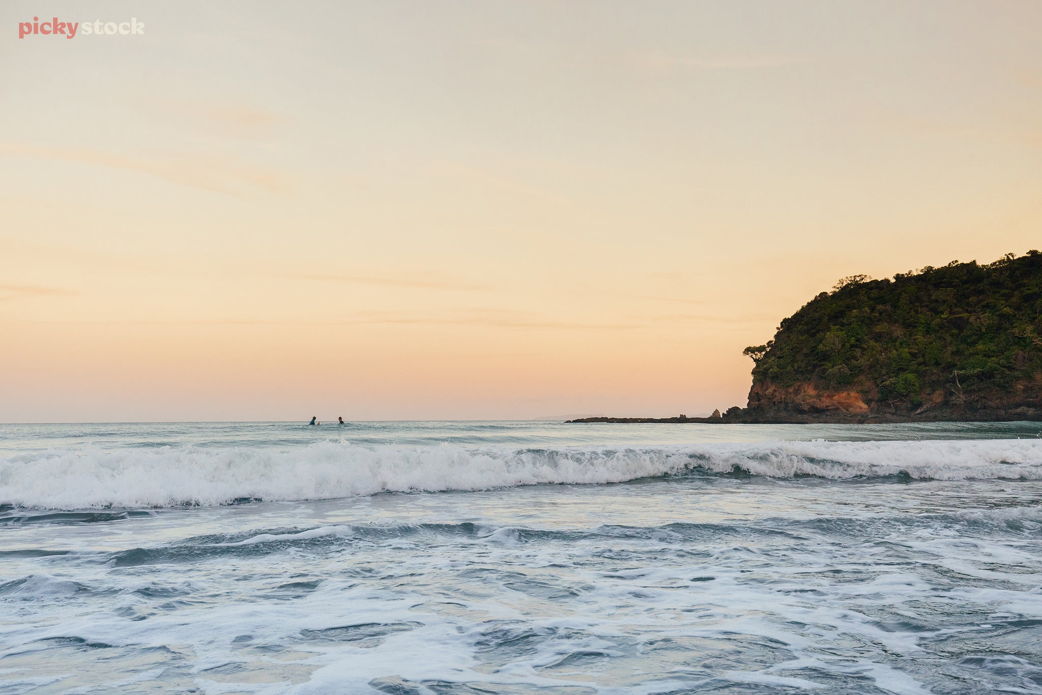 Landscape of two surfers waiting for a wave while small currents break against the shoreline under clear orange skies.