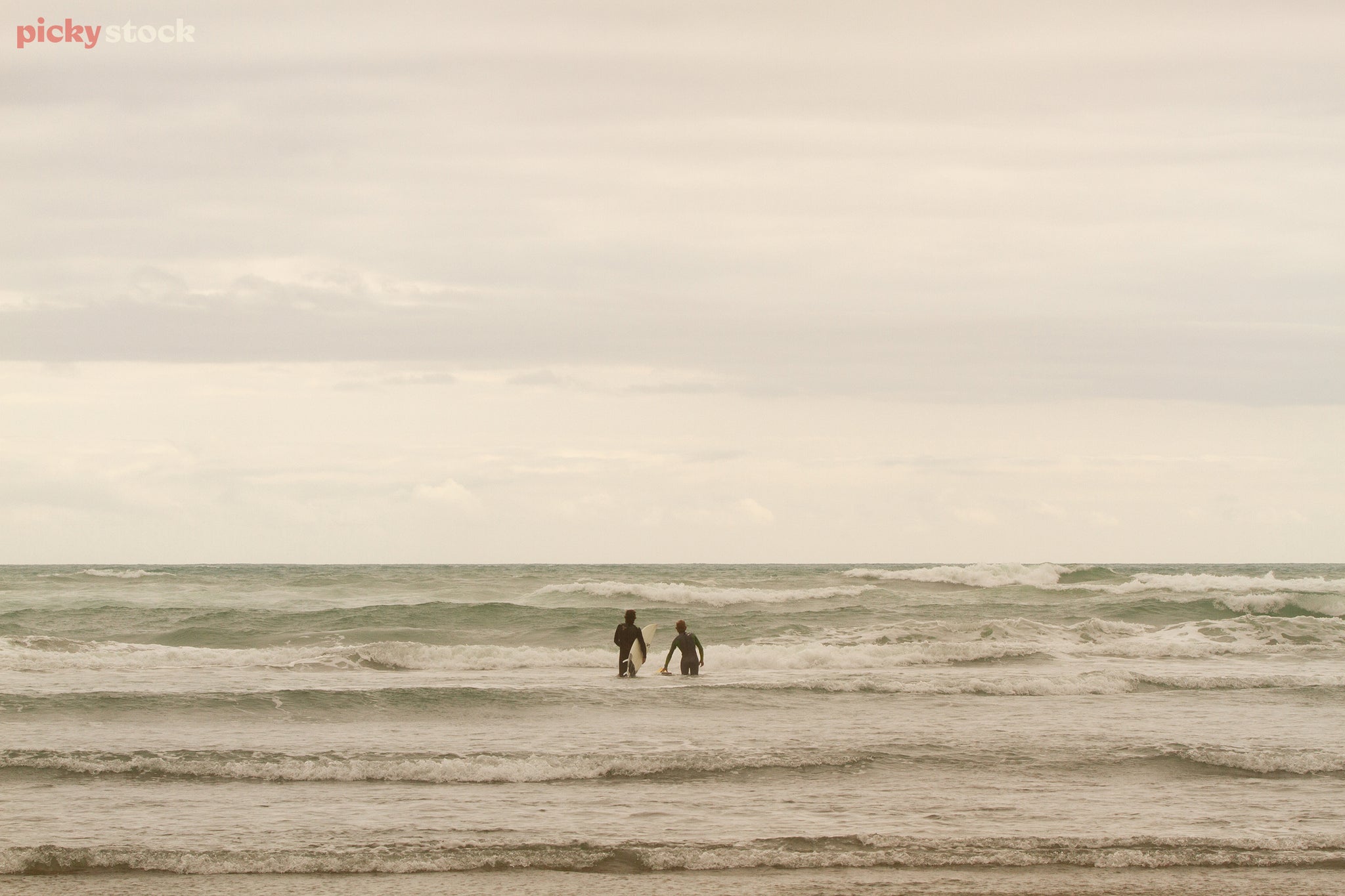 Landscape of two surfers in black wetsuits wading out into the ocean, the waves breaking against them under a clear bluish green sky.