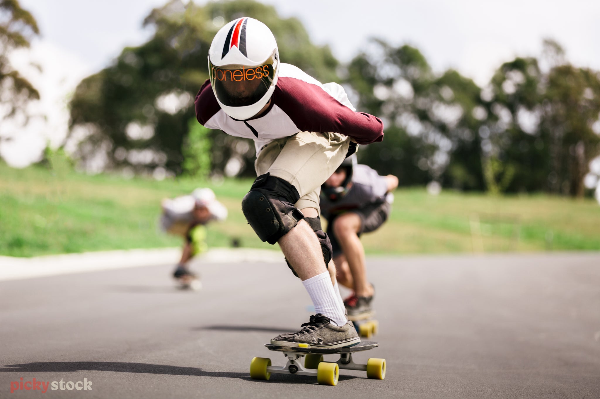 Landscape close-up of a group of longboarders speeding down a slope, the foremost is in black safety pads on their knees and a large white helmet with ‘boneless’ written across the visor.
