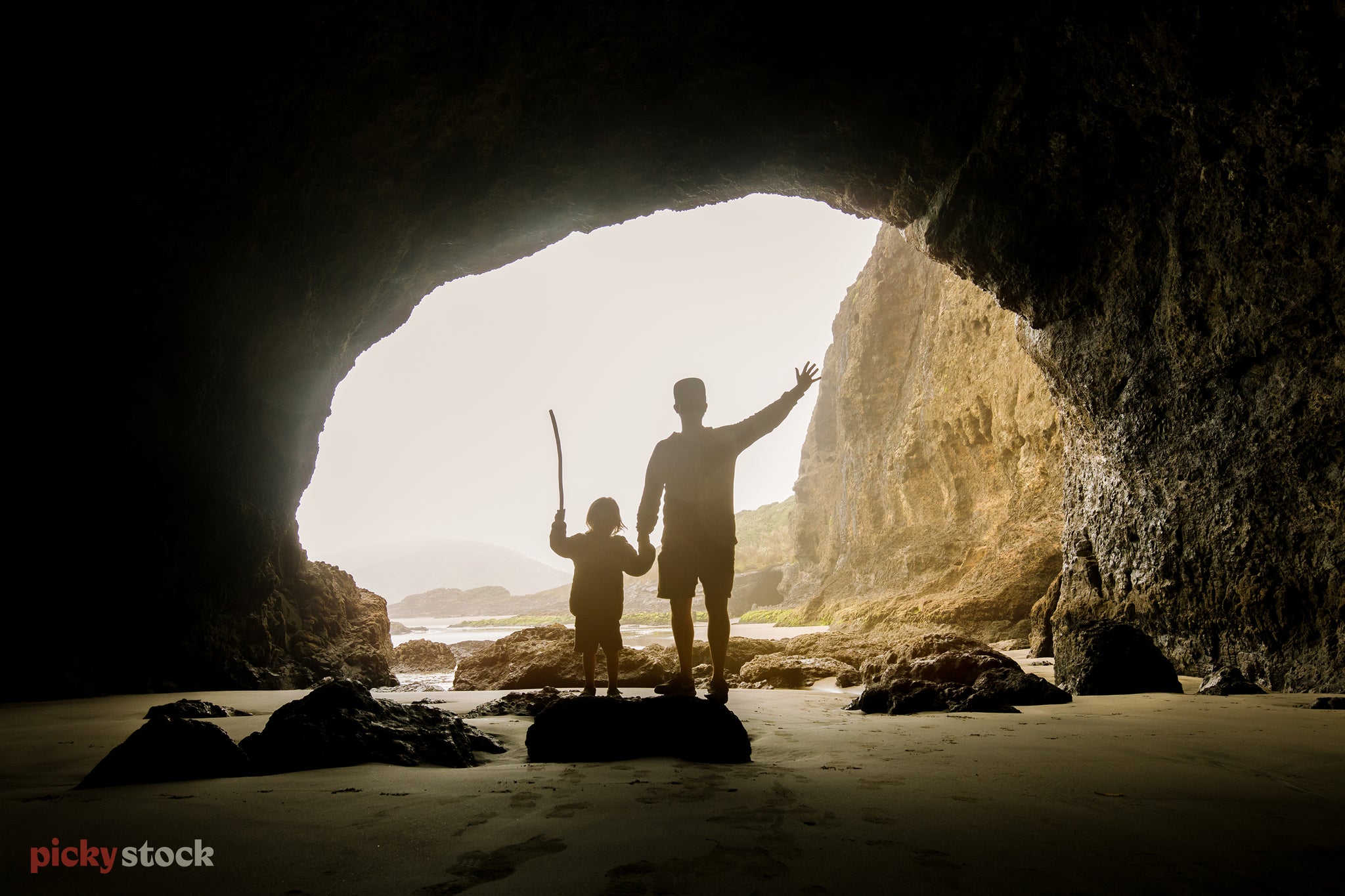 Landscape of an adult and child silhouetted in a beachside cave. The child waves a stick high above their head atop a rock.