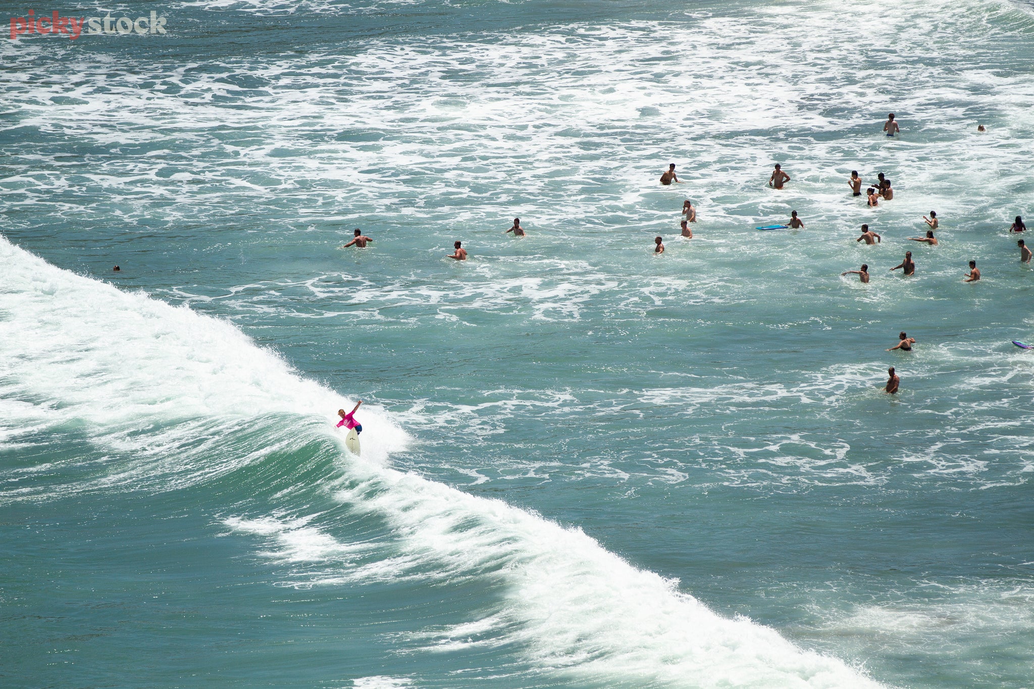 Landscape of female surfer in pink gliding over a crashing wave while two dozen beachgoers play in the clear waters nearby.