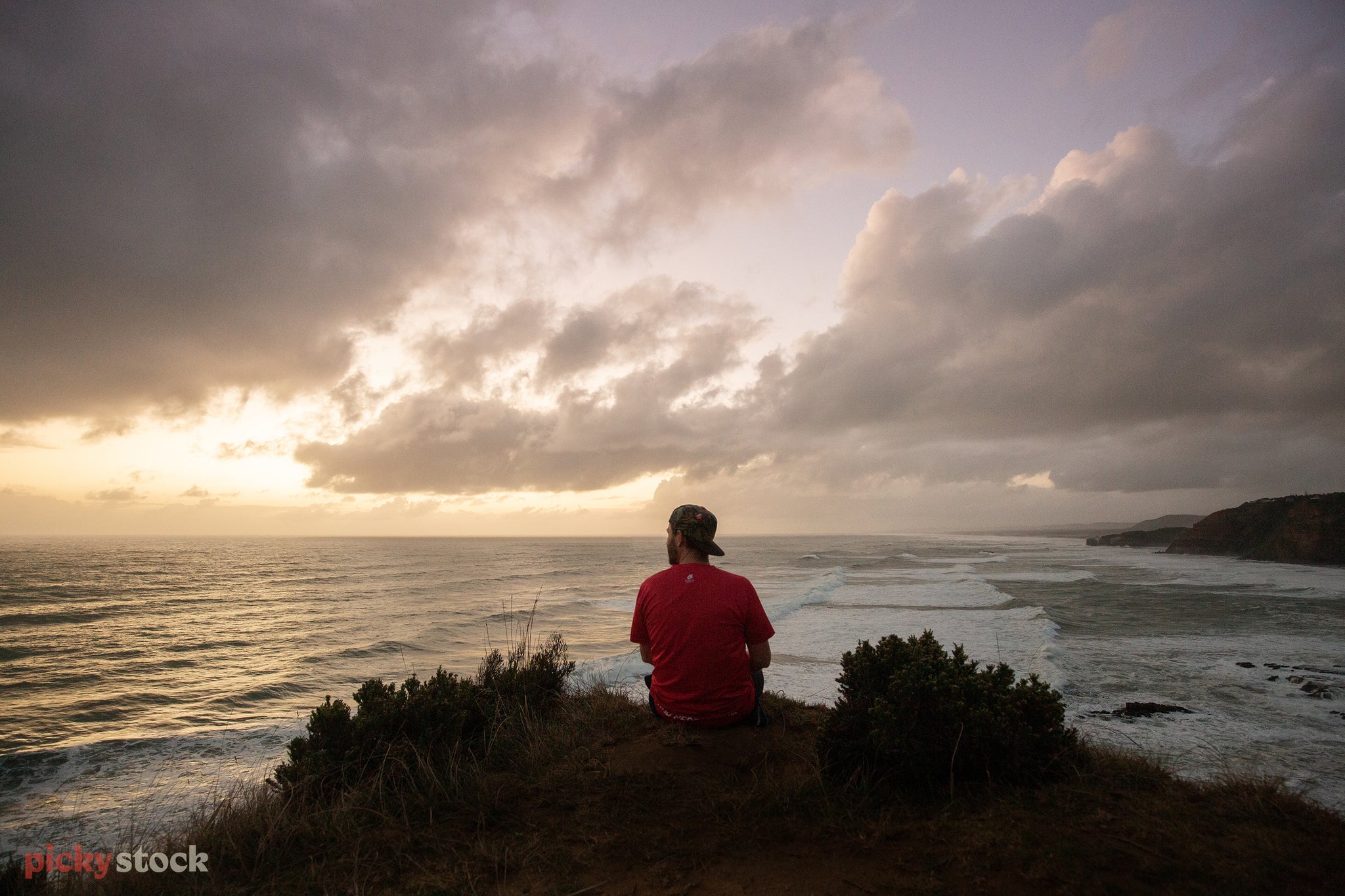 Landscape of a man in red shorts atop a grassy hill over looking at the sunset as waves break along the shore.