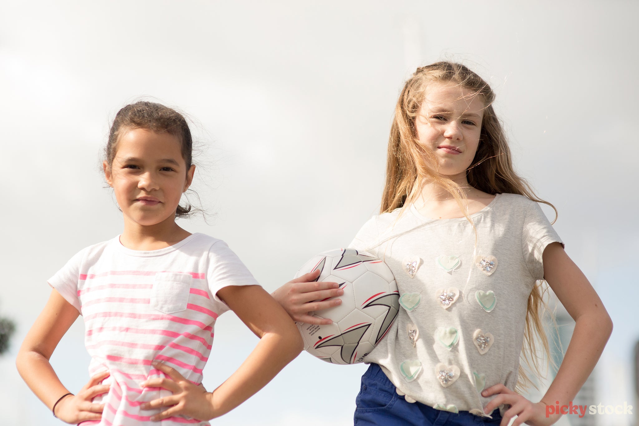 Landscape close-up of two young girls looking confidently to camera, hands on hips, one with ball in hand. 