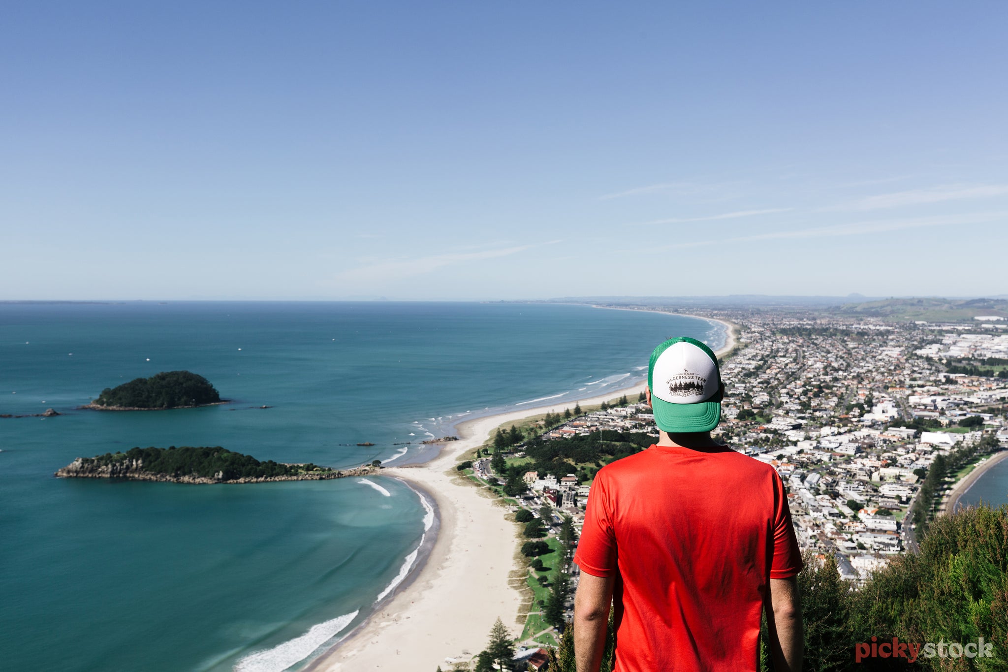 Man in red shirt with green and white hat looks out from the top of Mount Maunganui, down to the beach bellow on a bright blue day.