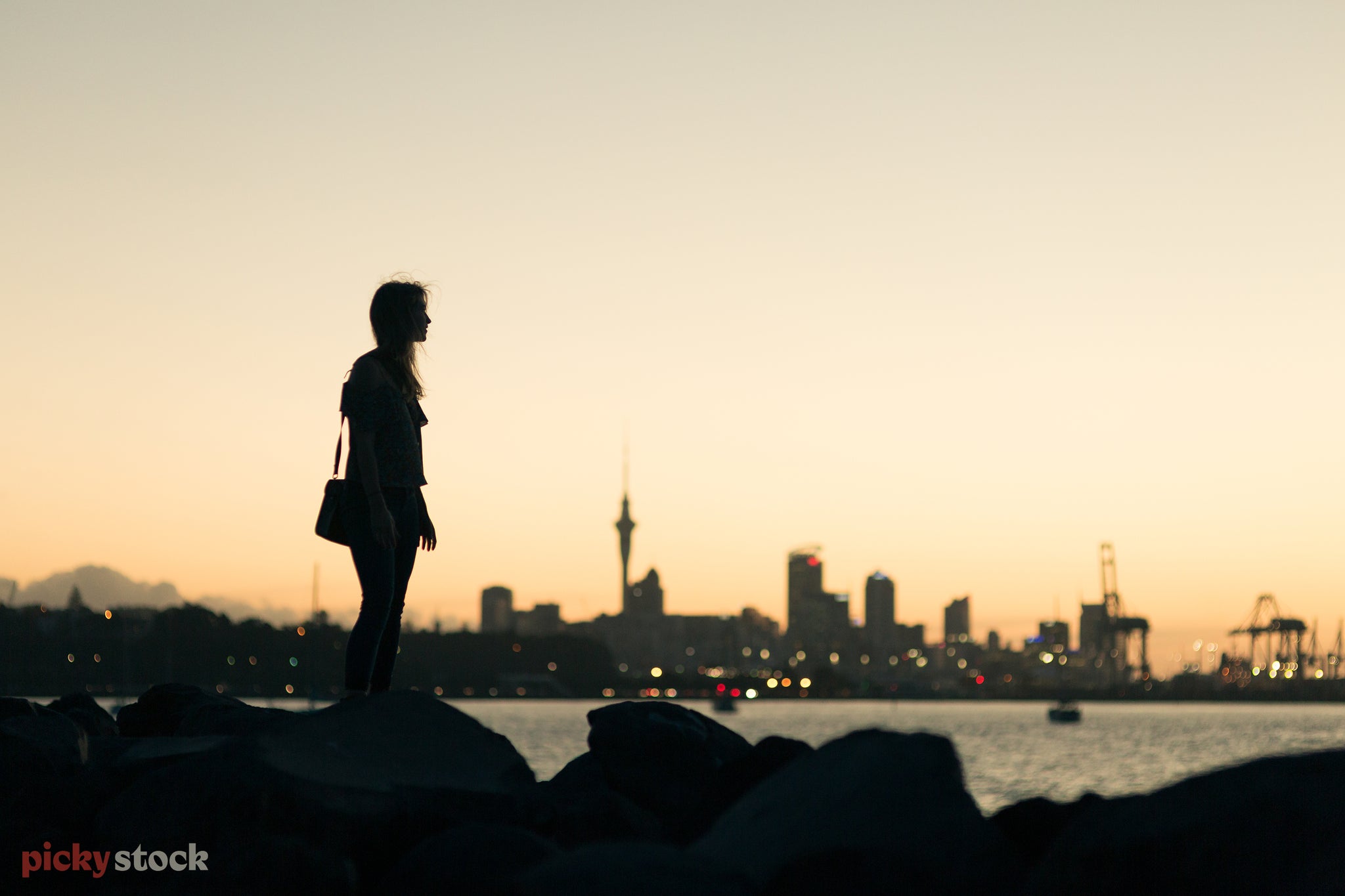 Silhouette of young female standing on rocks, in front of Auckland city. The light is dusk-like, and the city lights are just turned on. 