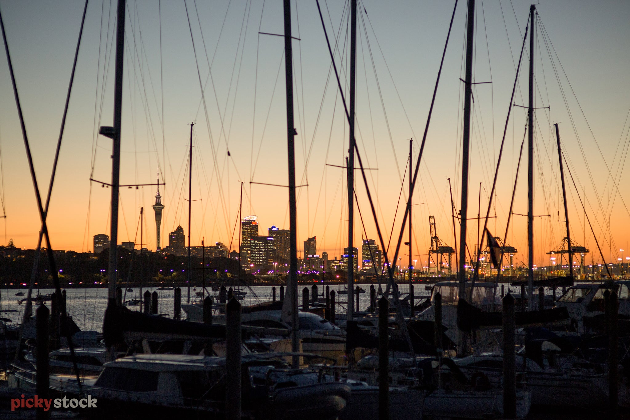 Looking through the silhouetted masts of sailboats, back towards Auckland City. The sun is setting on a perfect day, the city lights just turning on. 