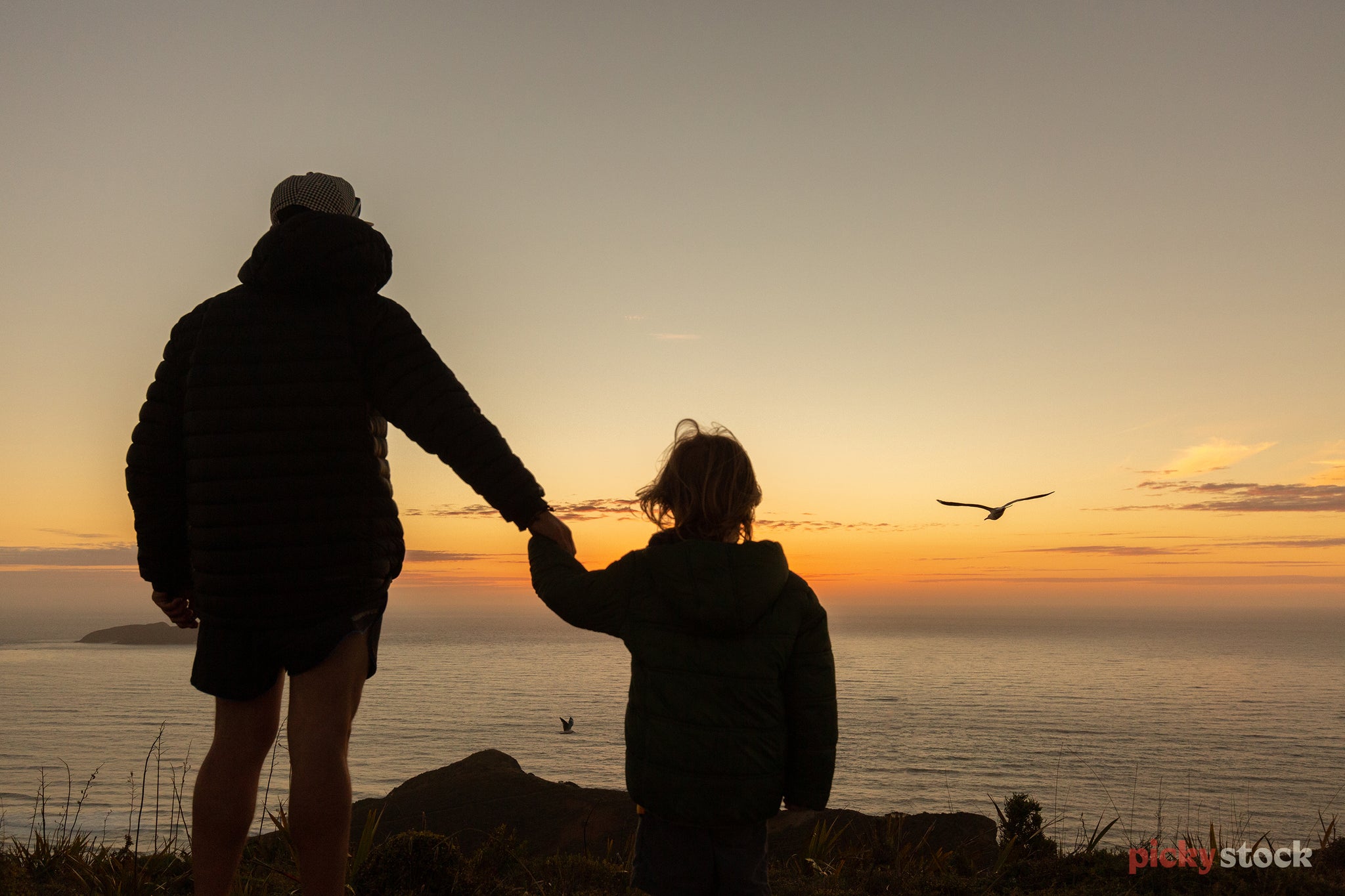 Silhouette of dad in puffer jacket holding hands of young daughter as they look out from a cliff towards the ocean as the sun sets. 