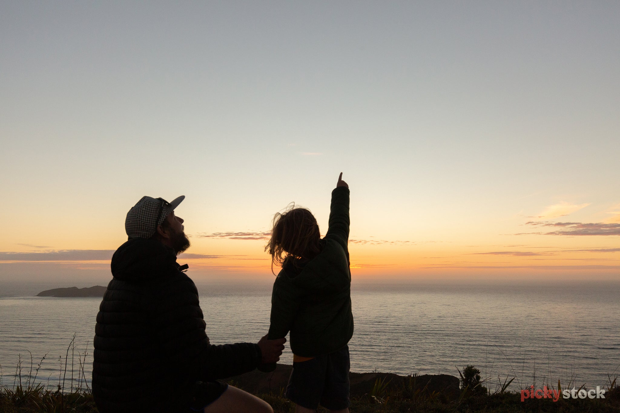Silhouette of dad in puffer jacket holding hands of young daughter as they look out from a cliff towards the ocean as the sun sets. The dad kneels down to her level, and she points up to the sky, spotting something. 