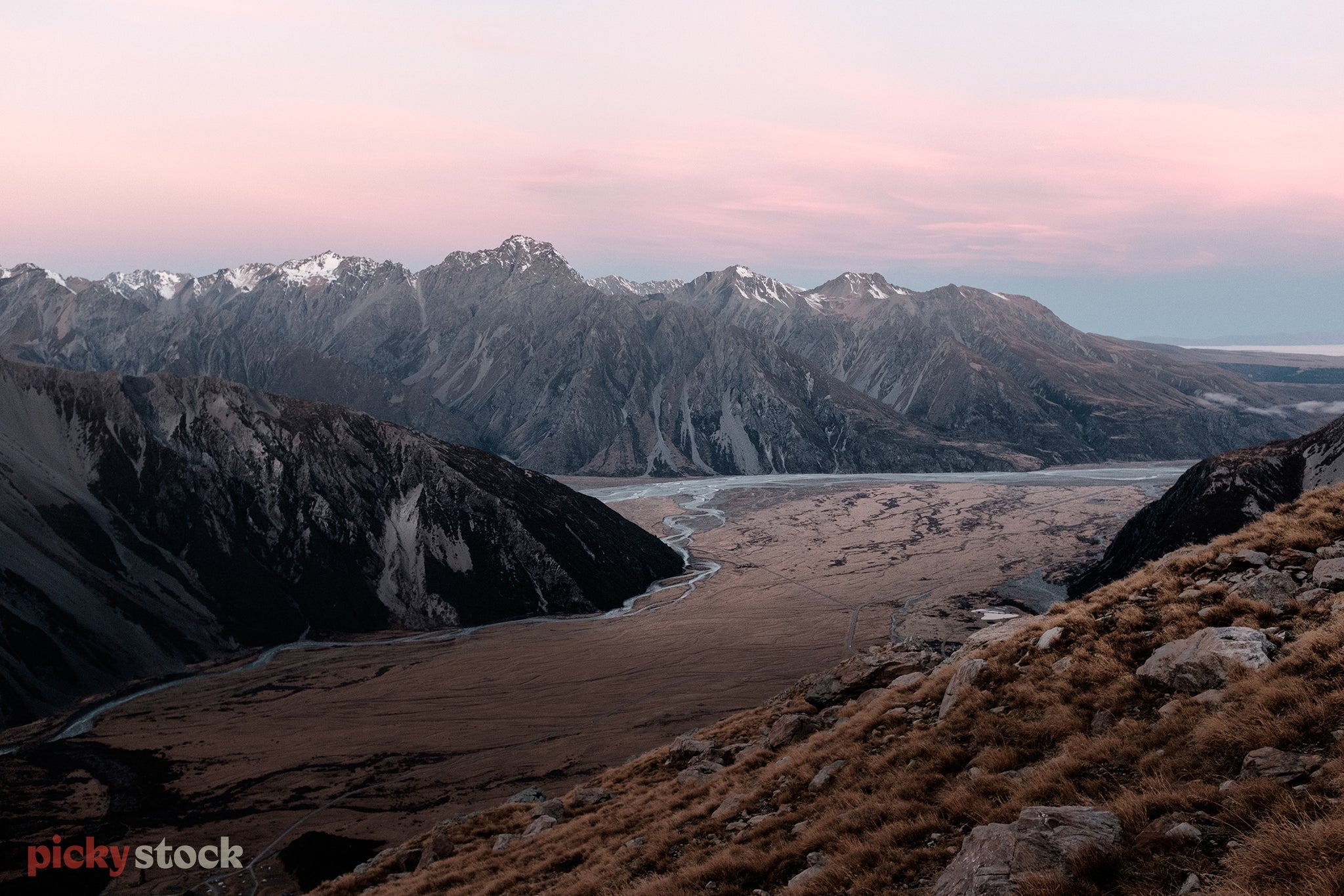 Looking out from a tussock ridge line towards the snow covered mountain peaks of the Hooker Valley. The light is turning pink as the sun is setting. 