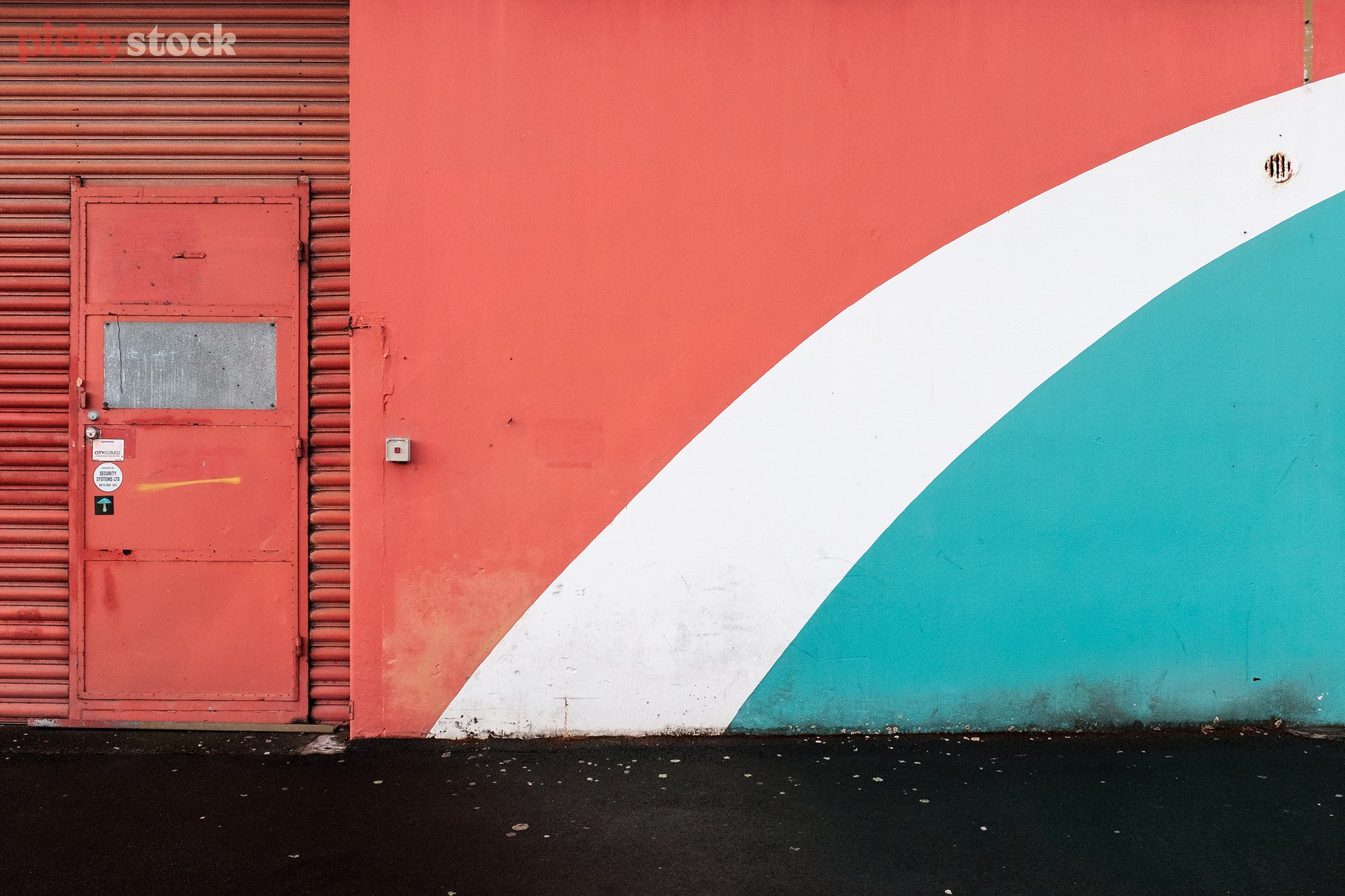 The side of a building, half corrugated iron, half concrete with a door to the left. The entire building is a faded painted red, with light white and mint green stripes painted to the bottom right. 