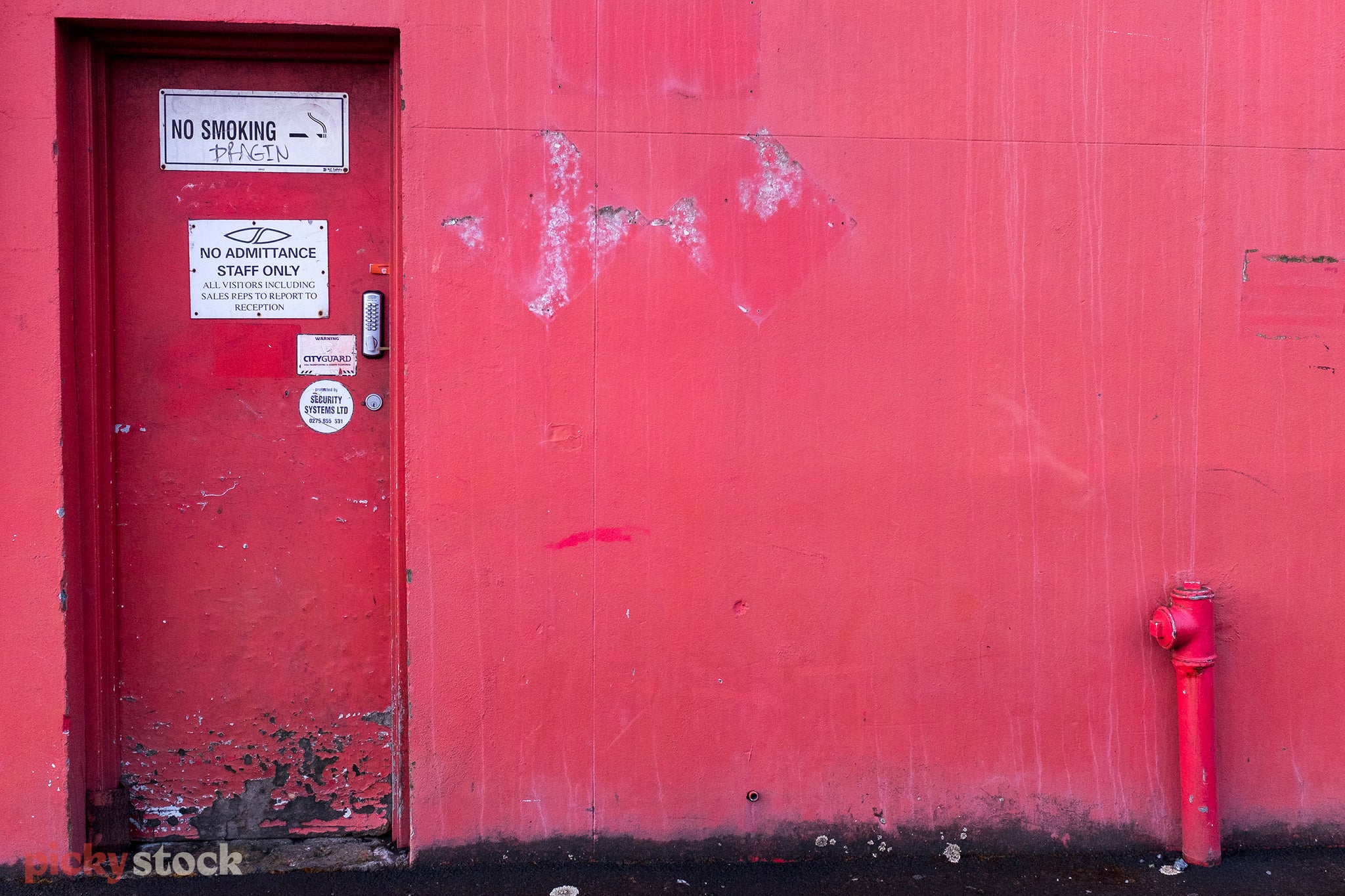 The side of a concrete building with a maintence door to the left. The entire building is a faded painted red. With a "no smoking" sign on the door that's been graffitied. 