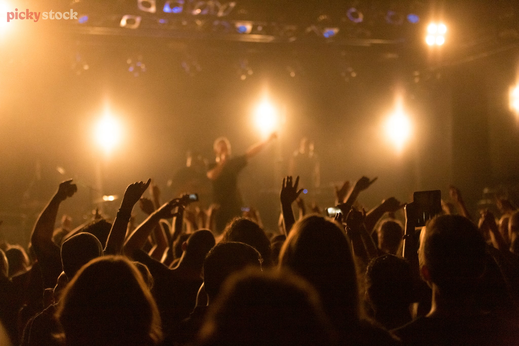 In a concert crowd mosh-pit, close to stage as hands and arms are raised in the air. Band is unrecognisable as the orange lights block the viewpoint. 