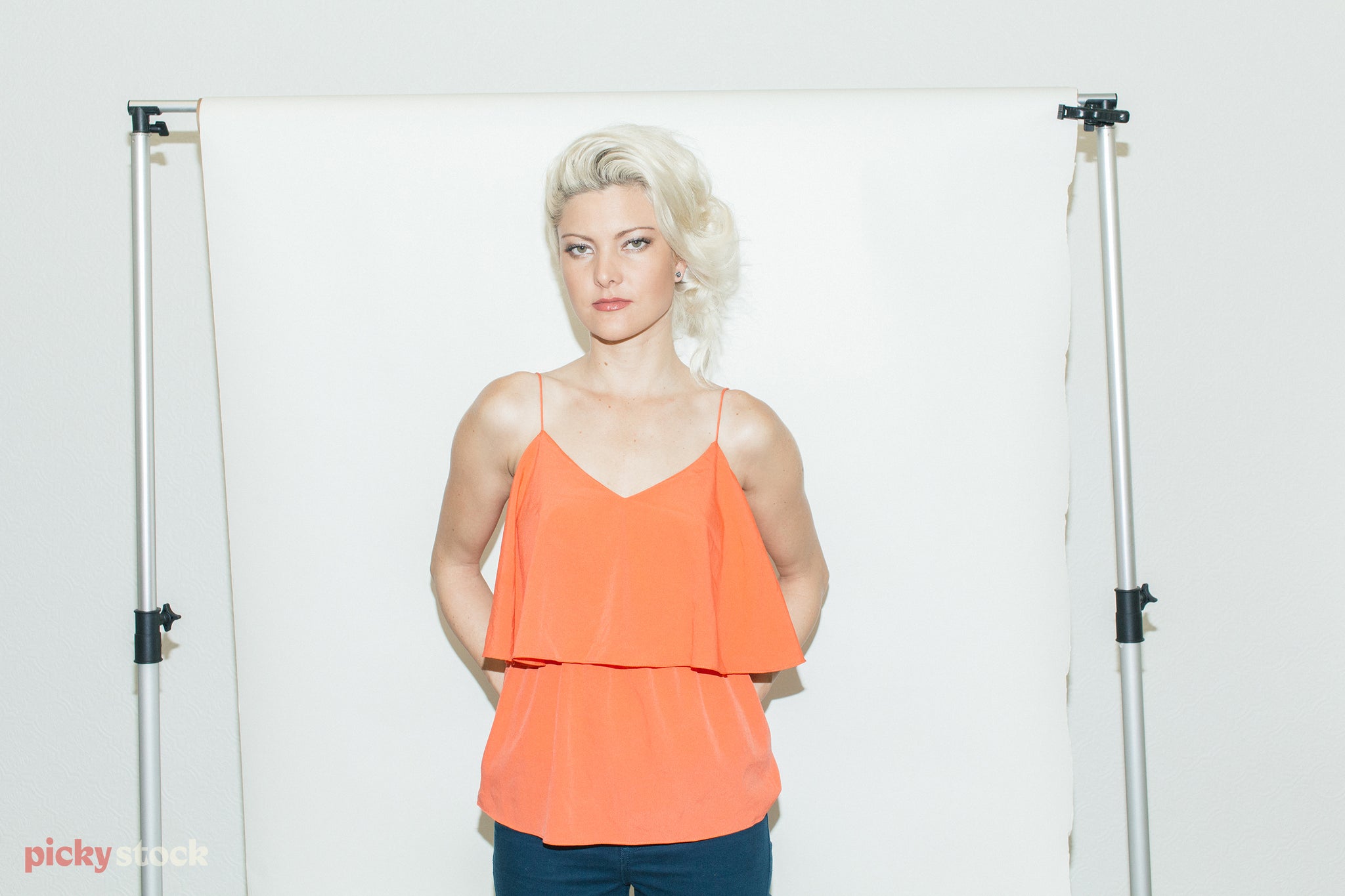 Lady with blonde Marilyn Munroe hair looks direct to camera. She wears a thin strapped orange top, and stands in front of a white photographic studio backdrop. 