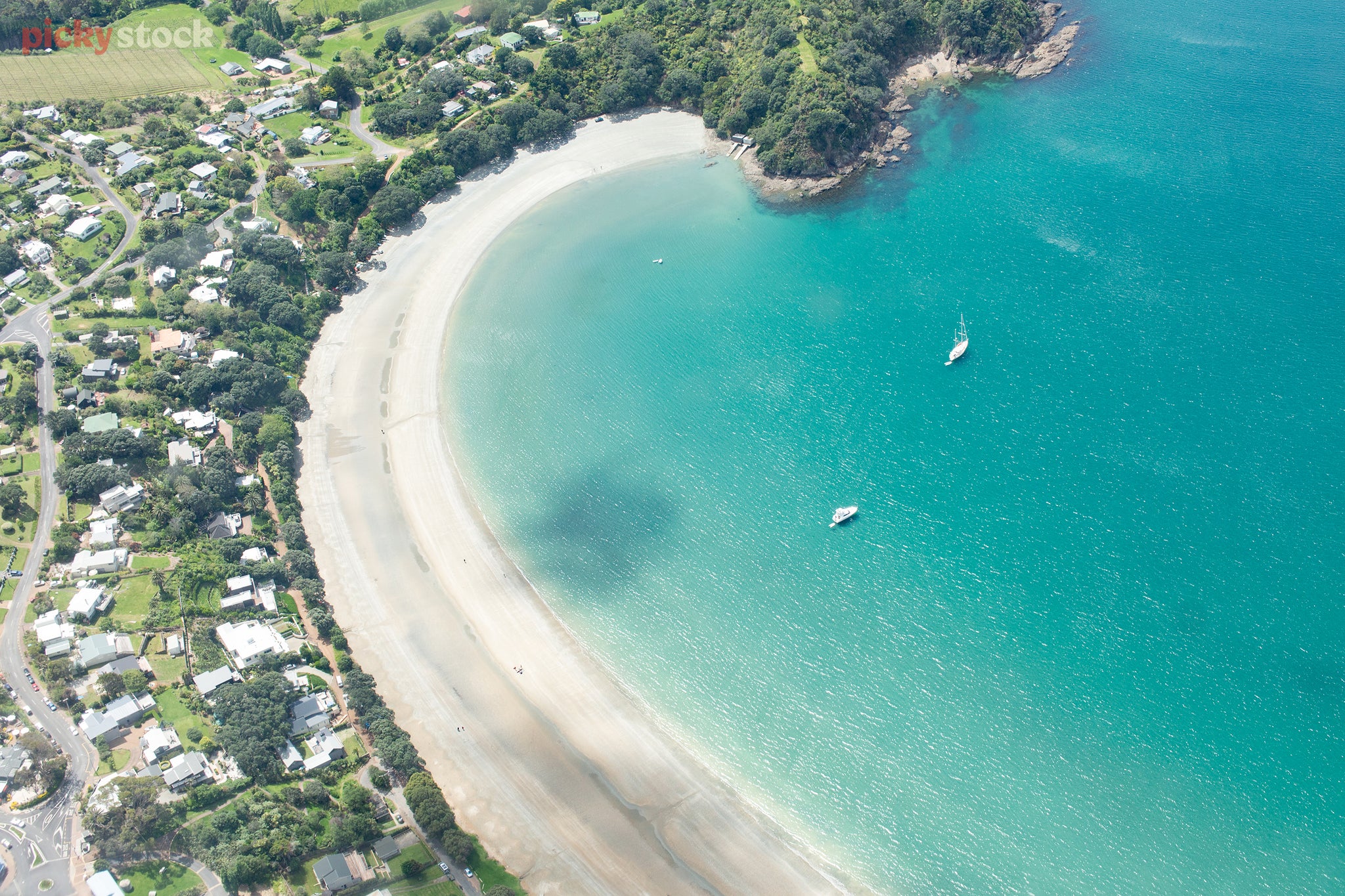 Aerial image of curved bay, where batches, sand and sea all meet. The water is vivid and turquoise with two white sailboats moored close to shore. 