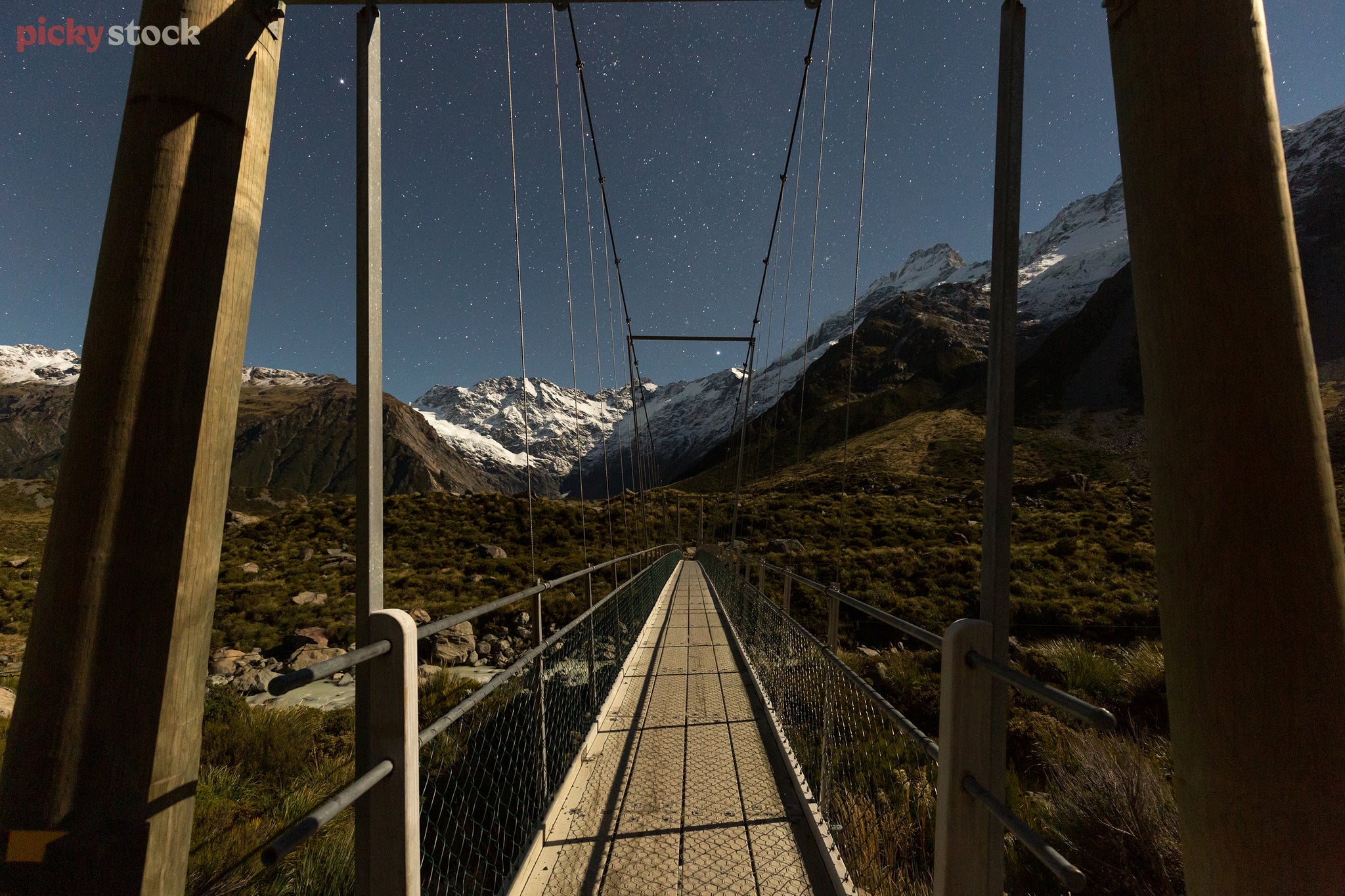 Crisp, high definition image looking over a very symmetrical hikers bridge towards the starry night sky. Snow-capped mountain range can be seen in the background. 