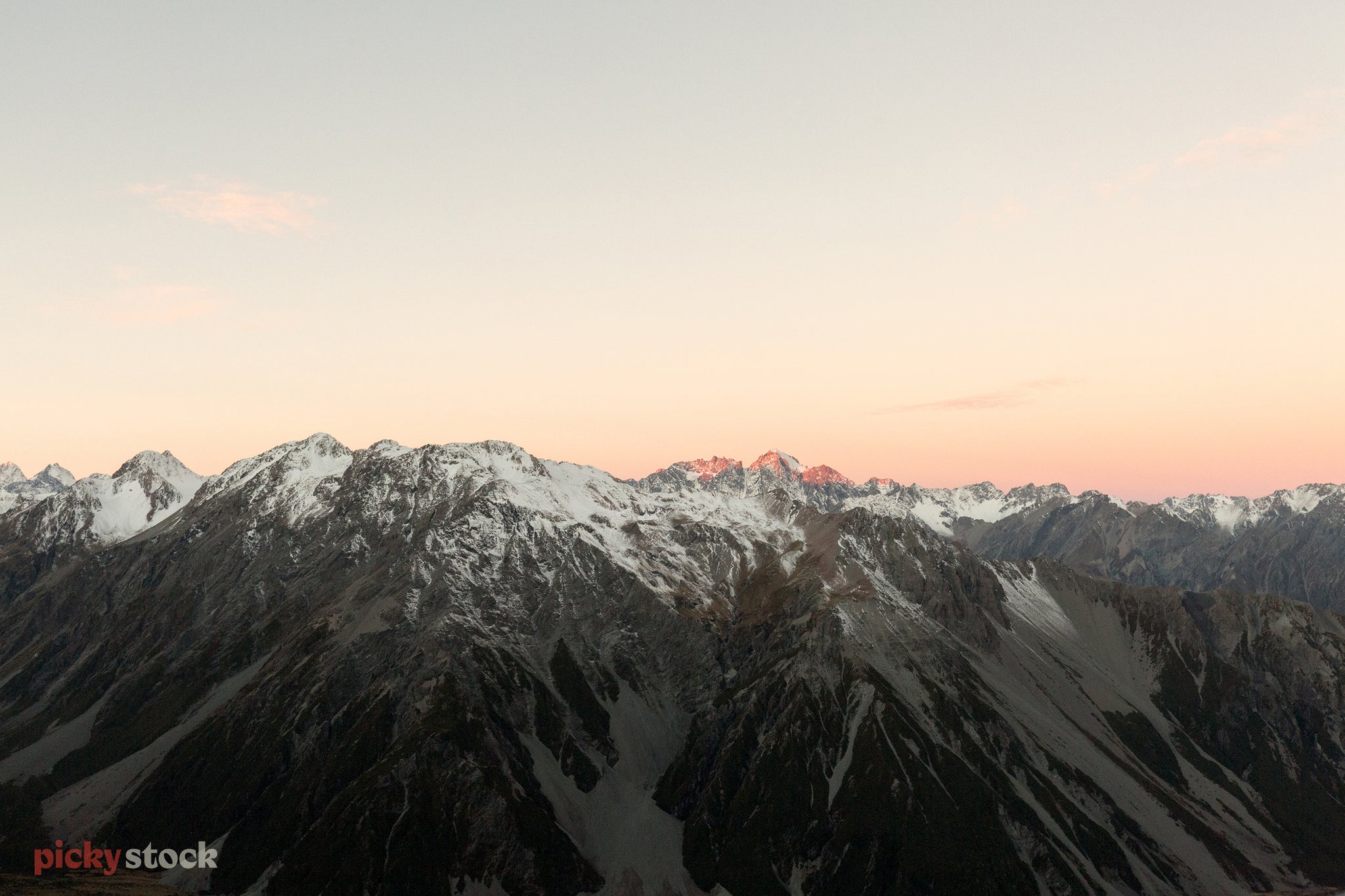 Looking out over the Mount Cook mountain range, just snow capped at the top, with the sheer mountain revealed beneath. The dusky pink light of sunset reflected in the snow. 