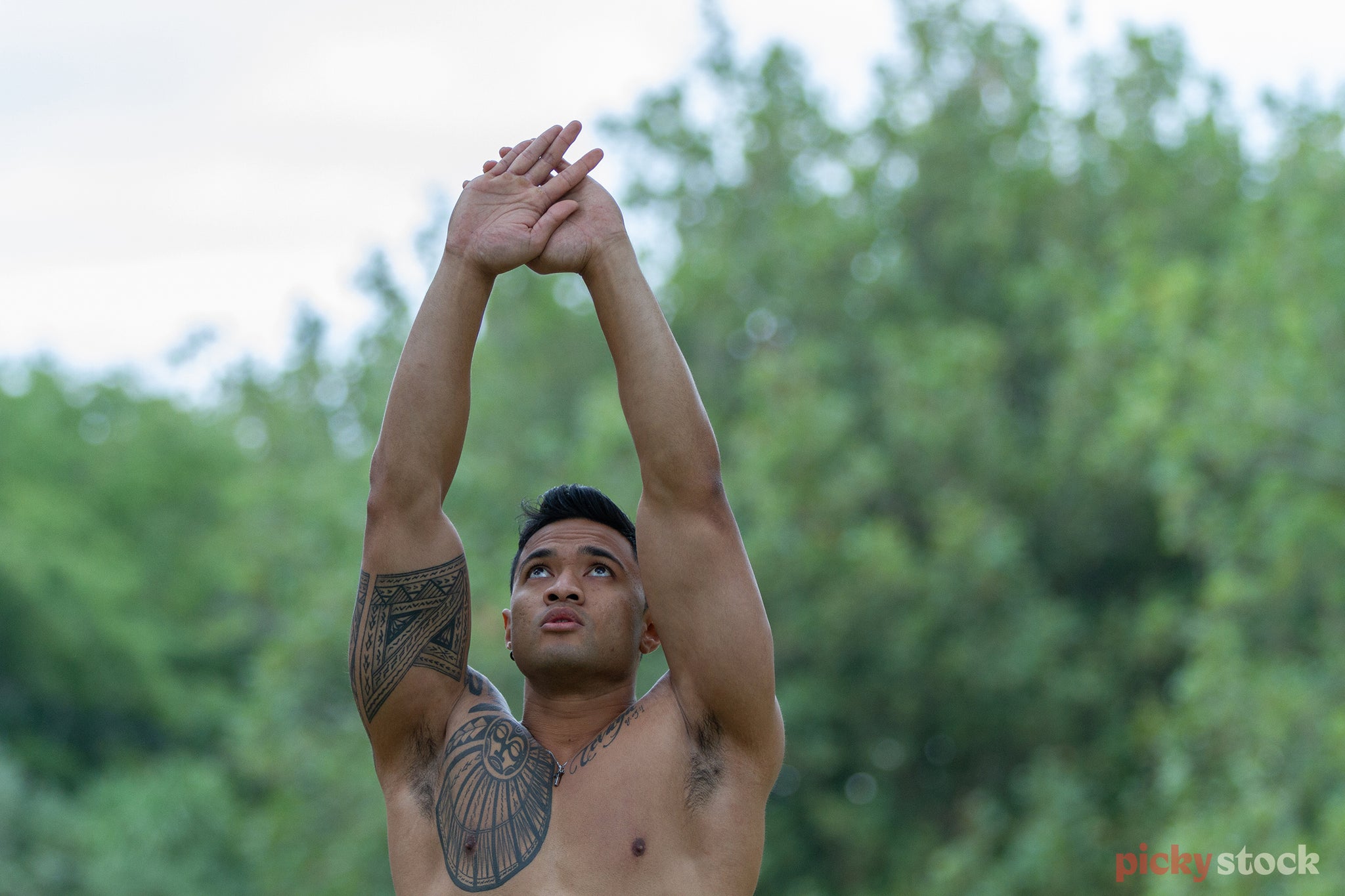 Polynesian man with arms outstretched above his head, gazes up towards his head in a moment of exerice. Shirtless, his traditional tattoos can be seen on his upper right chest, shoulder and wrapping around his bicep. He stands in what looks to be a park, which is out of focus. 