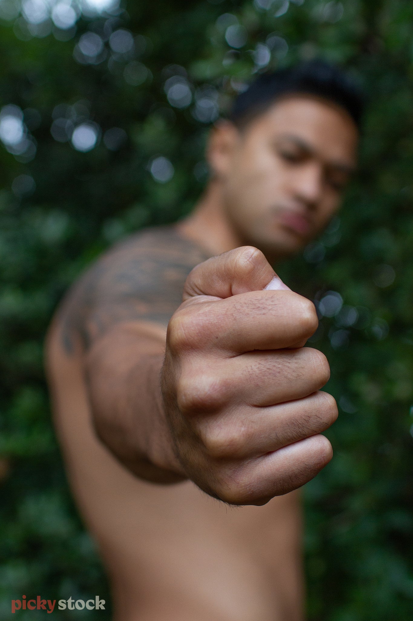 Polynesian man holds his fist out to camera. Fist in focus, everything else not in focus as we can see he is shirtless, his traditional tattoos can be seen on his upper right chest, shoulder and wrapping around his bicep. 