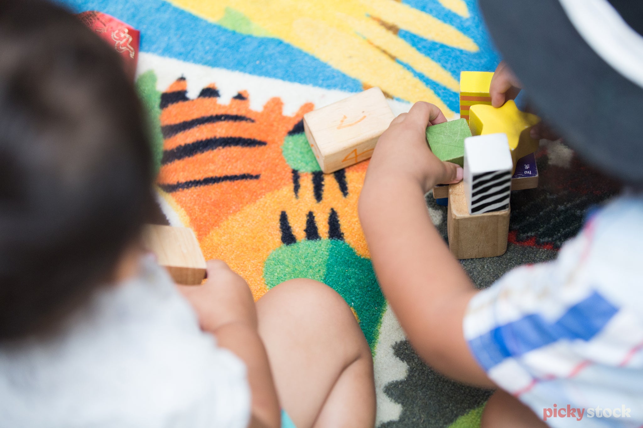 Two toddlers play with wooden building blocks on the bright primary-coloured matt at pre-school. The children are unrecognisable as we see over their shoulders. 