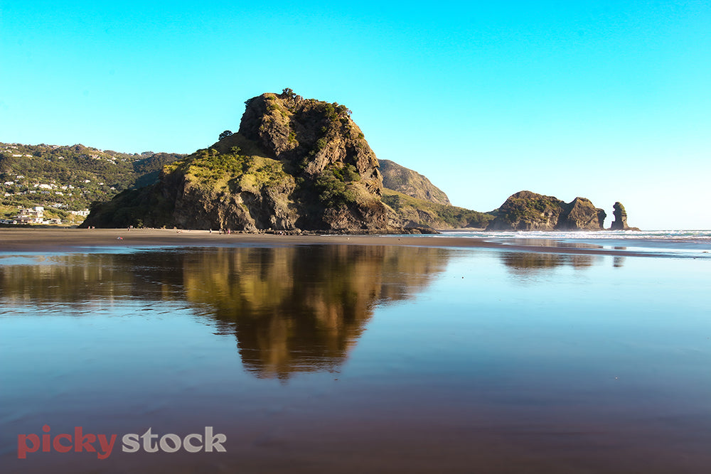 Reflection of Lion Rock, against the water & black sand of Piha beach on a blue day.