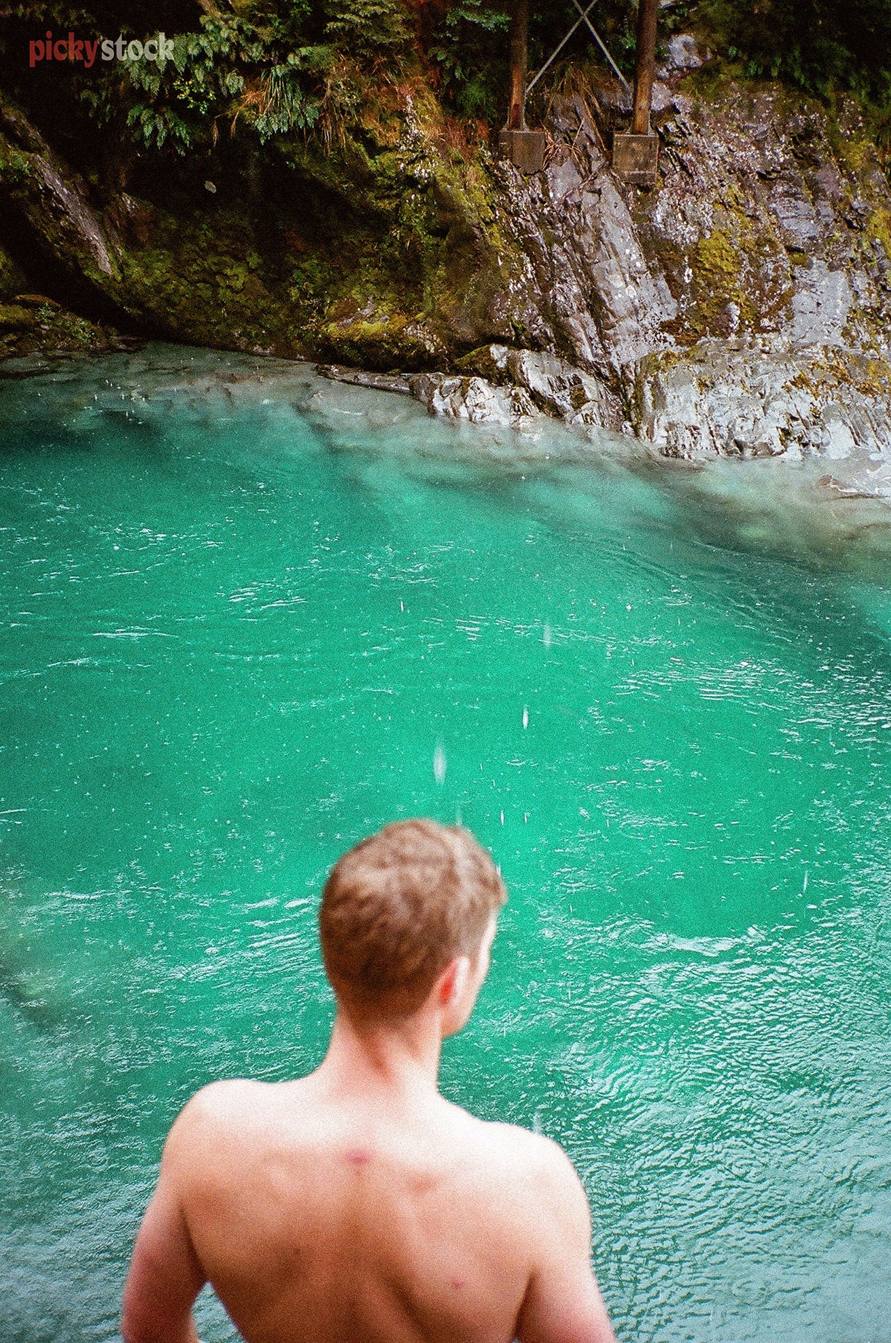 High angle filmic image of a young man looking out to emerald blue cold water. He looks to have been in the water already, and is ready to jump back in. 