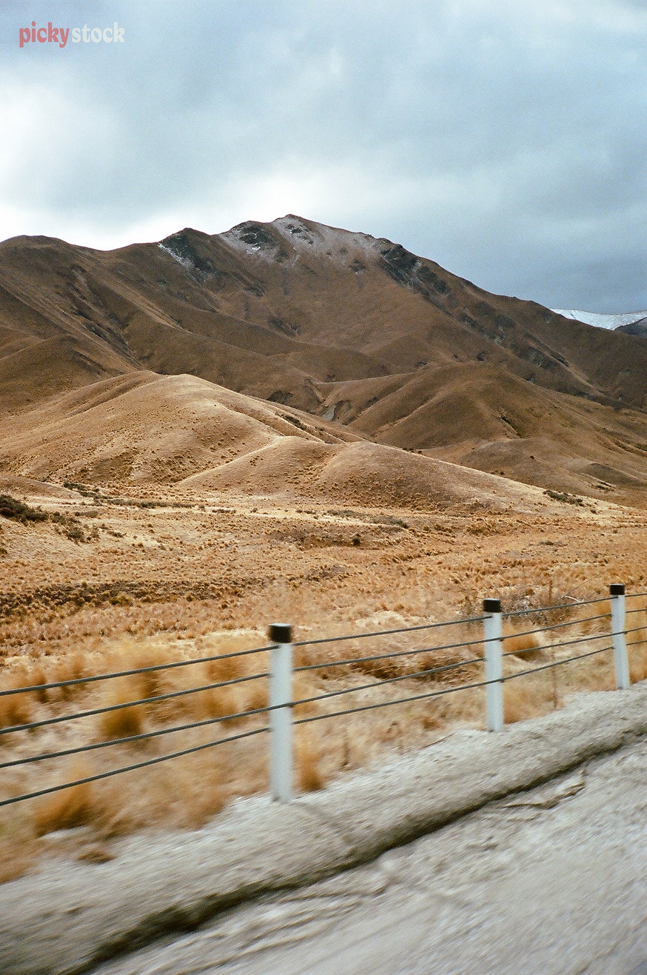 The rolling sepia-coloured hills of the Lindis Pass as seen from a car moving at speed. We see the side of the road and the road barriers before the hills.