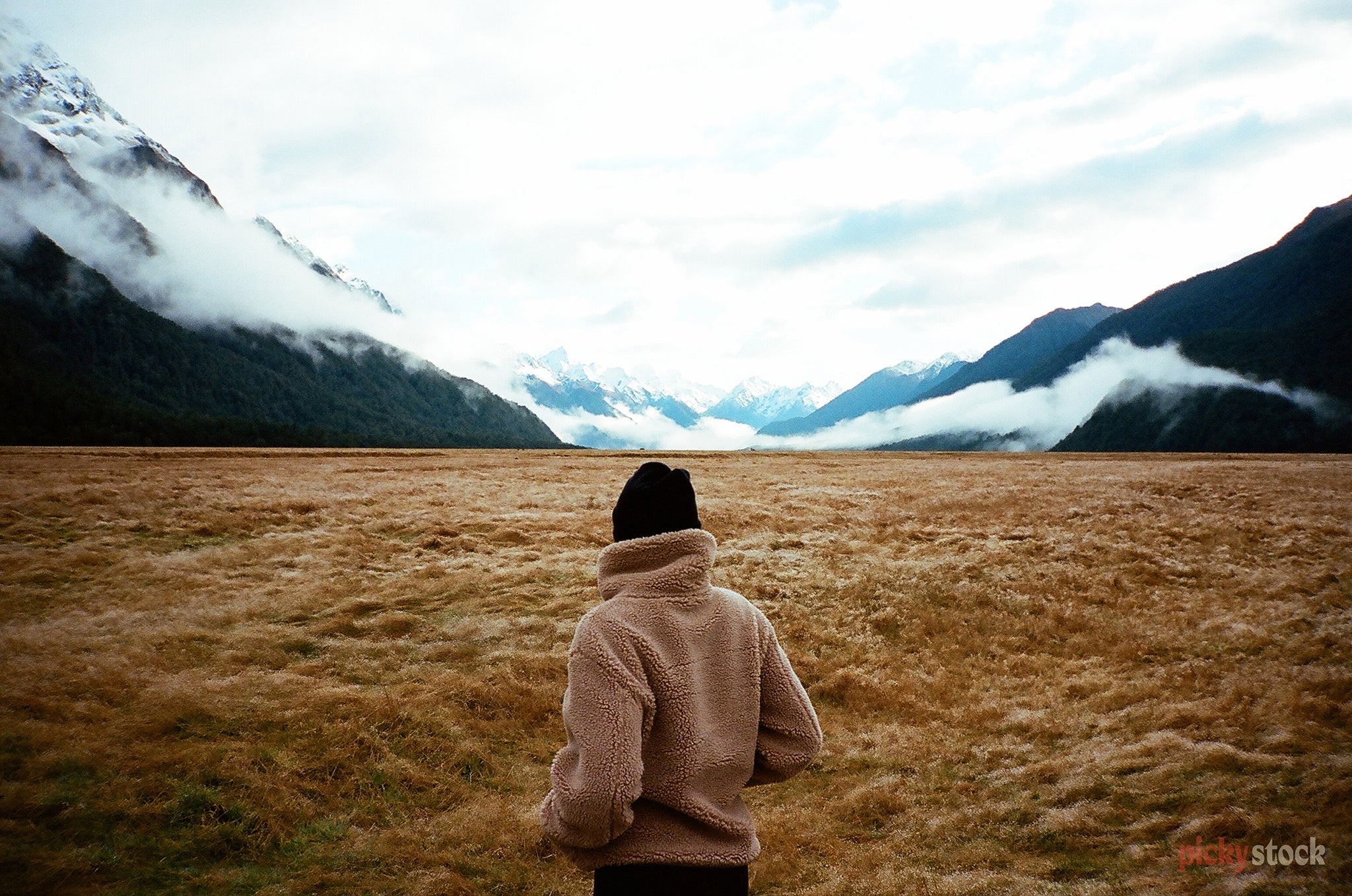 A lady in a jacket looks out across the fields of the Eglinton Valley, Milford Sound, out towards the ice-capped mountains. There's low lying fog at the foot of the hill.