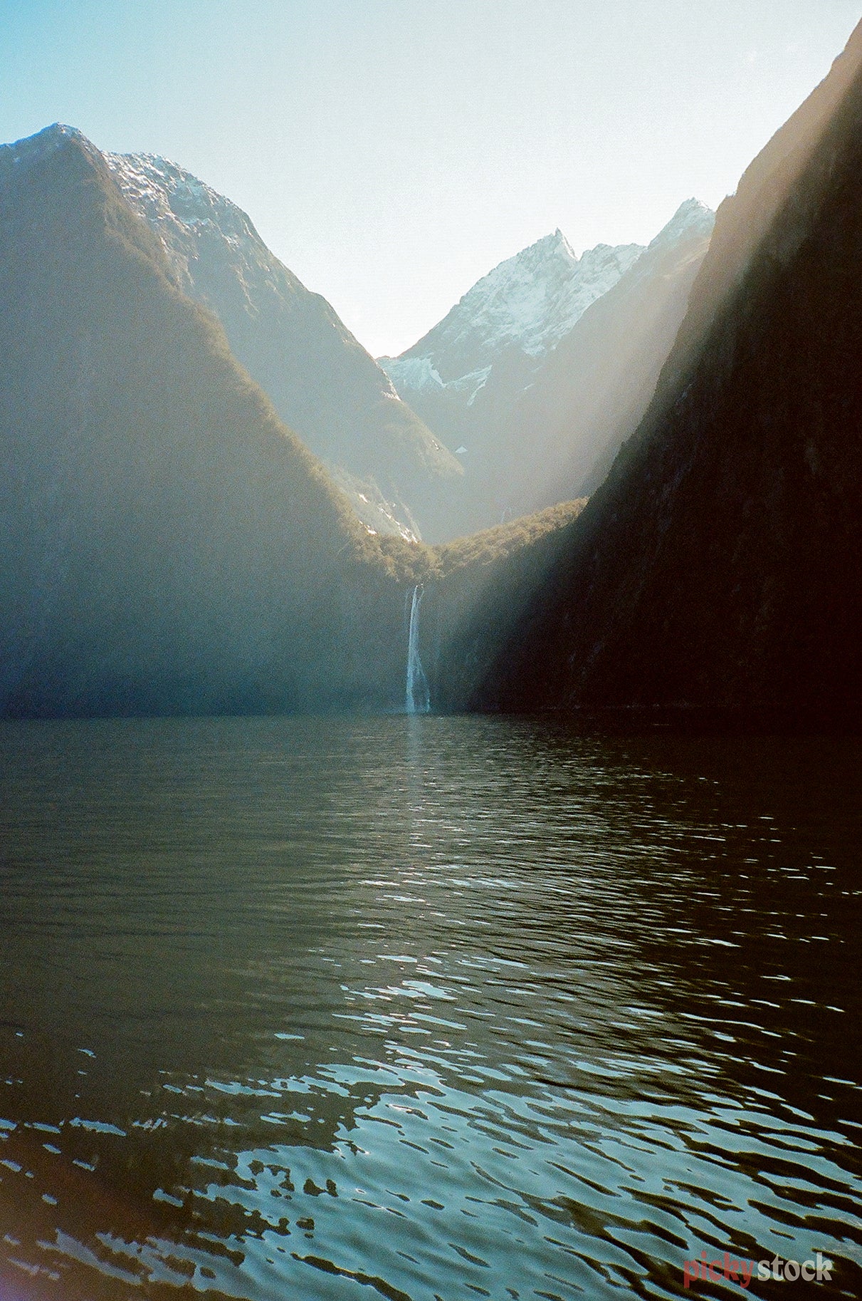 Looking out across the waters of the Milford Sound, towards a small waterfall at the foot of mountains. Sunlight filters in through the valley hitting the water. 