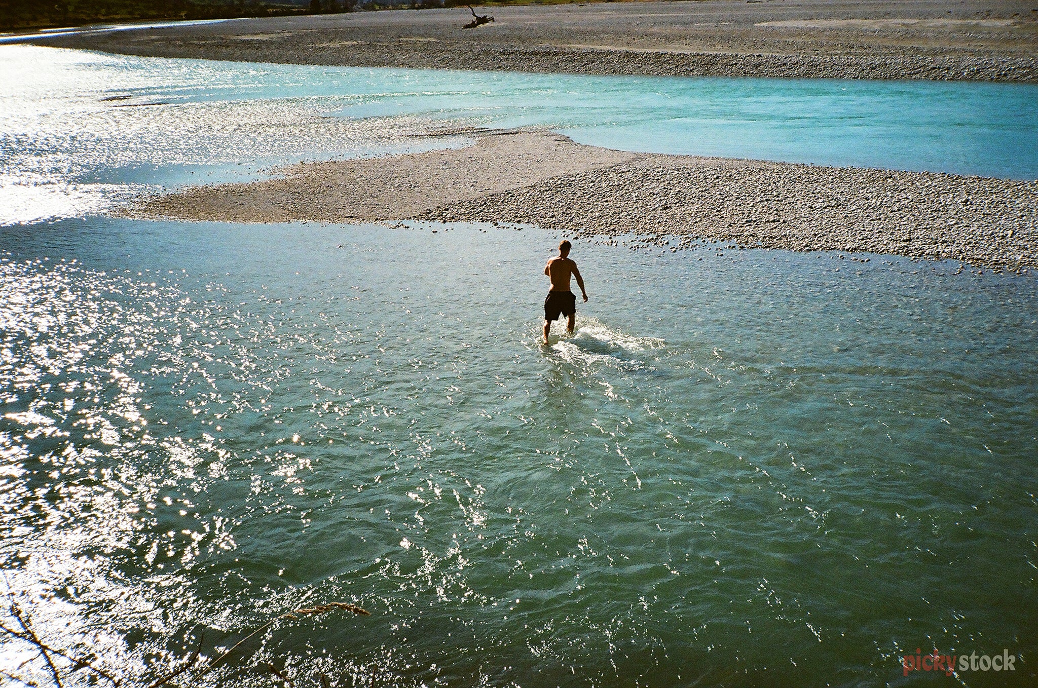 A man dashes out of the cool blue water while swimming in Glenorchy, Otago. The camera takes the photo from a high angle at a distance.