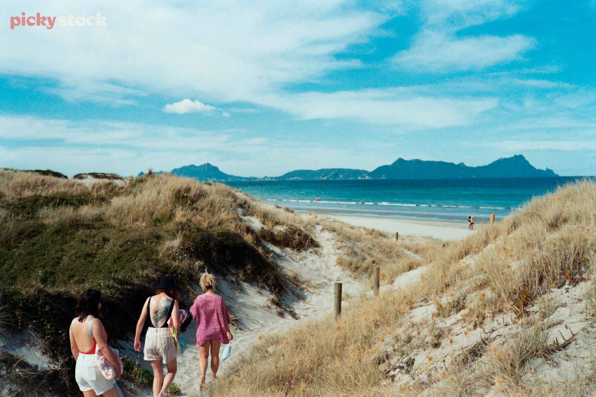 Looking out from the dunes out to see at Urititi beach on a blue summery day. Three young adult female ladies head down the sandy path towards the beach, their backs to camera. We see the large back tattoo of one of the ladies. 