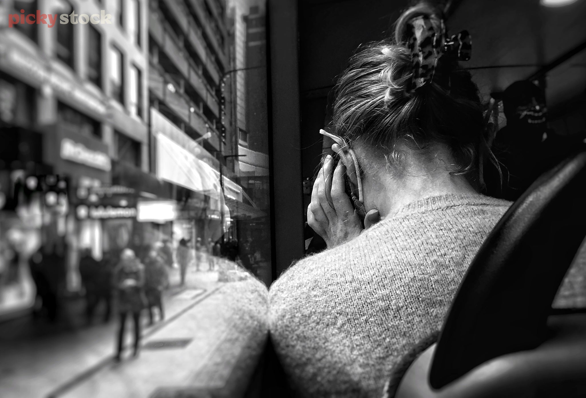 Black and white observational image from the back of a public bus. From over the sea, we see a lady at the window seat, hunched over on phone call, her hair clipped up in a bun. 