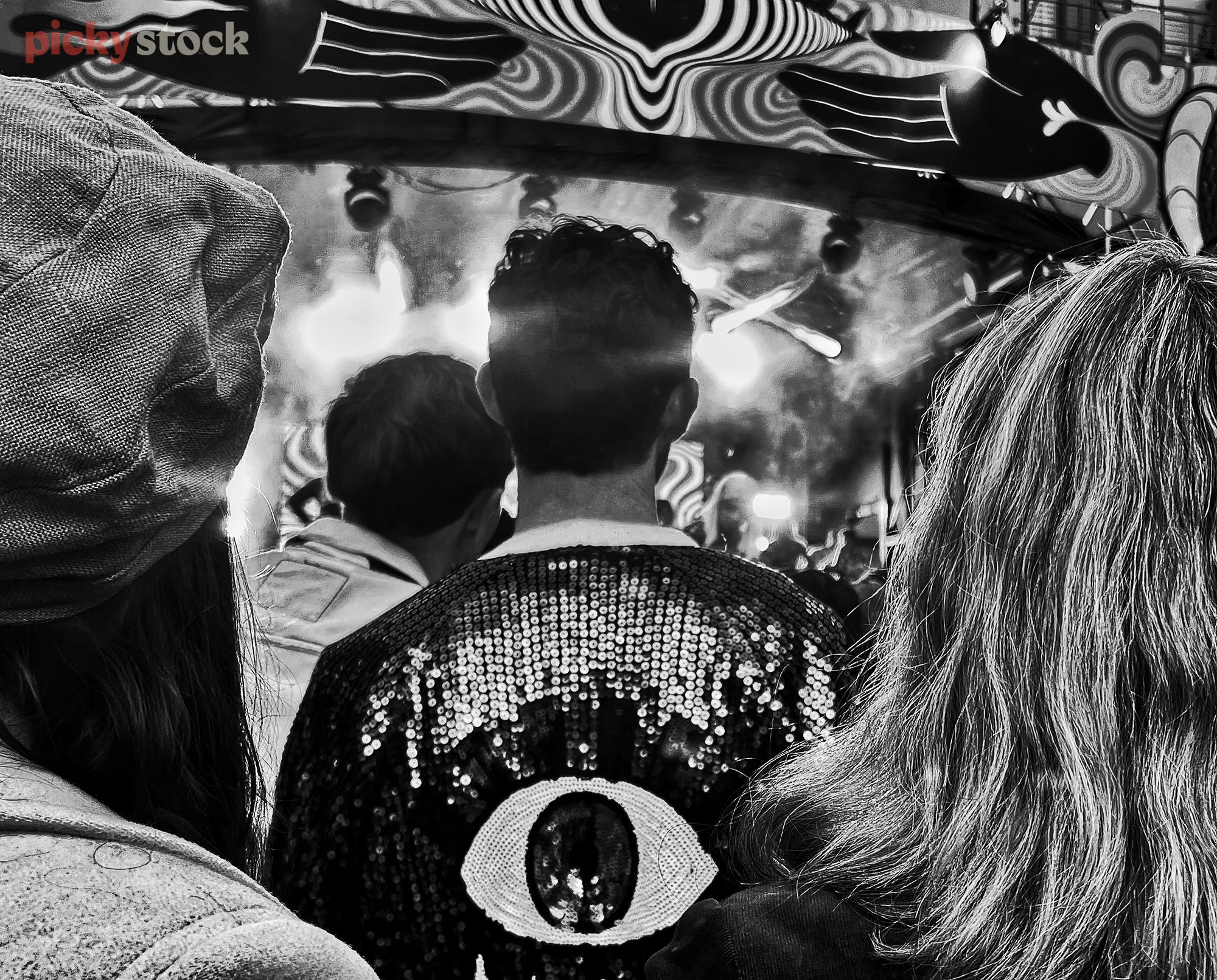 Black and white observational image in a music crowd. The viewer looks over the shoulders of a male and a female, towards a tall youn male with a sequined black jacket with a graphic eye on it. 