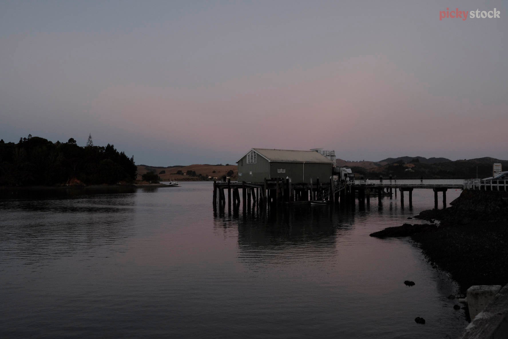 An old boating shed sits on a wharf, as the sun sets. The surrounding is still, misty and paint-like. Beautiful light pink reflections of the sky are reflected in the water. 