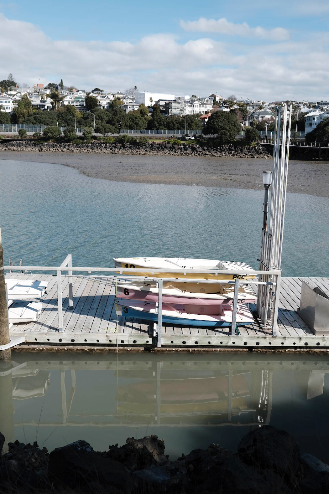 Looking down to a dock at Westhaven Marina in Auckland. On the dock there are 3 small pastel coloured beginners yachts stacked on top of each other – blue, yellow, pink. The colouring of the picture is undersaturated, so the blue sky and browny marina water are a bit dull. 