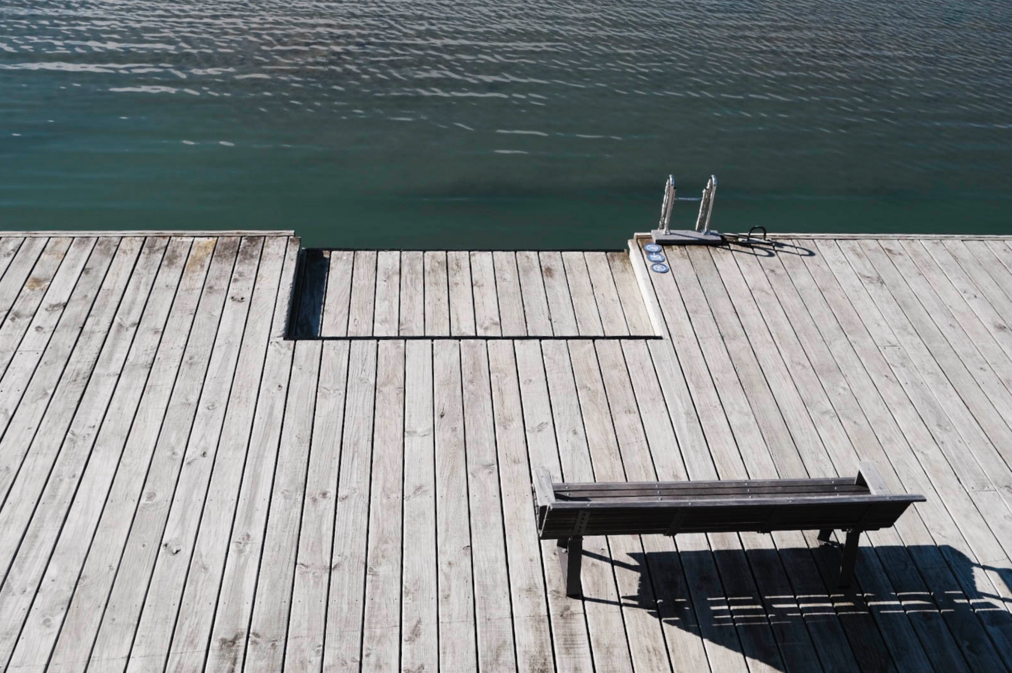 Looking down to a boardwalk at Westhaven Marina. The boardwalk cuts directly across the frame from left to right. There is water in the top half of the image. There sits a picnic bench in the bottom right of the image, with a small ladder to access the harbour from the boardwalk in the top right.