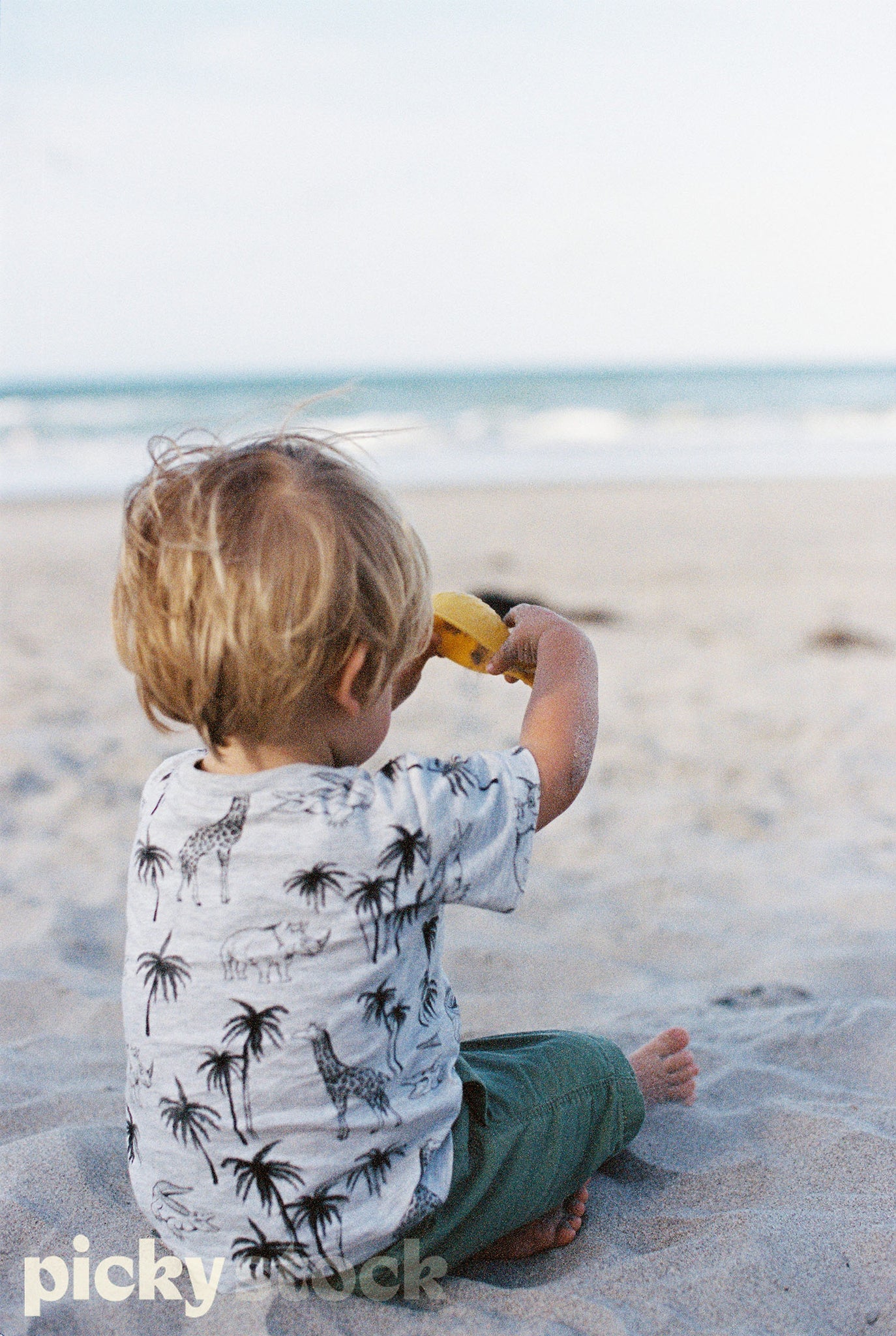 Young boy sitting on golden sand, holding a yellow spade. Wearing green shorts, with a grey top with a print of palm tree and giraffe. Ocean in background. 