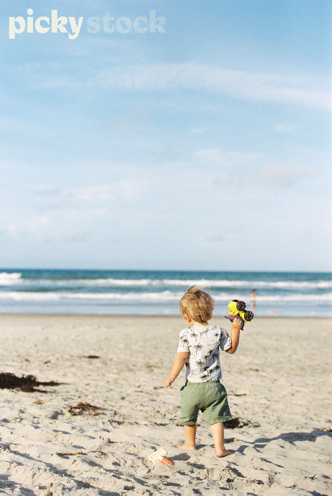 Portrait image of young blonde boy walking along golden sand beach. Holding small truck digger in right hand. Blue ocean with waves, and soft blue sky. Boy wearing green shorts with grey top