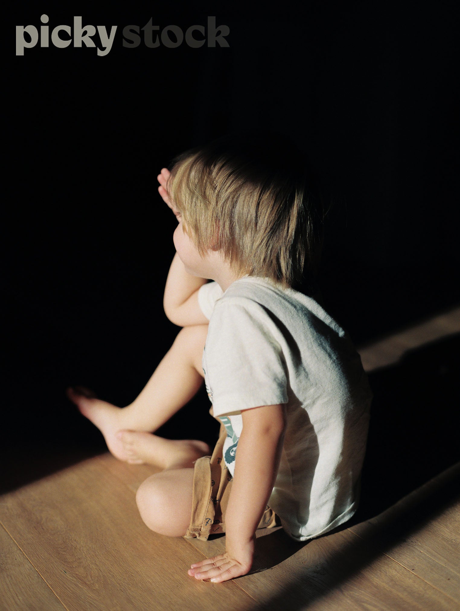 Small blonde young boy sitting on wooden floor. Sitting in light, with dark shadows surrounding him. 
