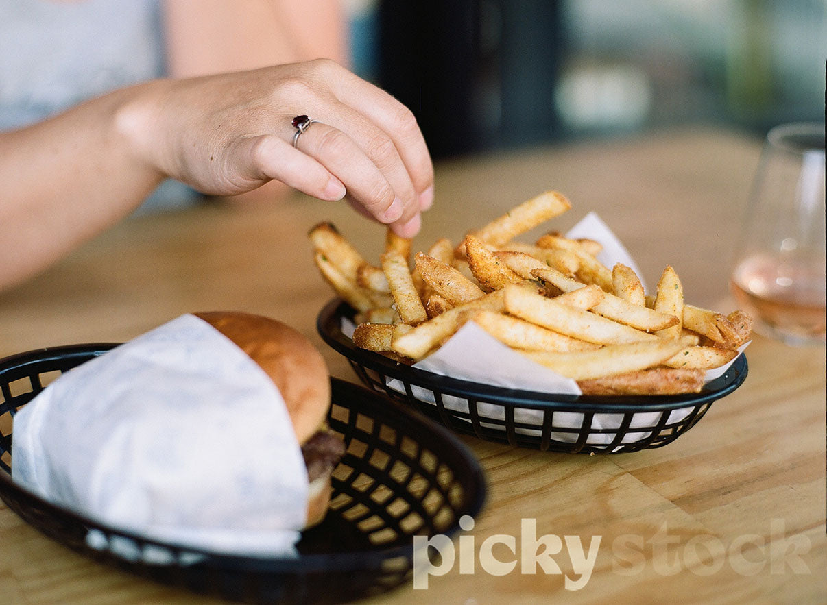 Lady with right on ring finger picking up a hot chip from a basket of fries. To the right of her is a cheese burger, wrapped in paper also sitting in a black basket. Food is sitting on wooden table with a wine glass on the right of frame