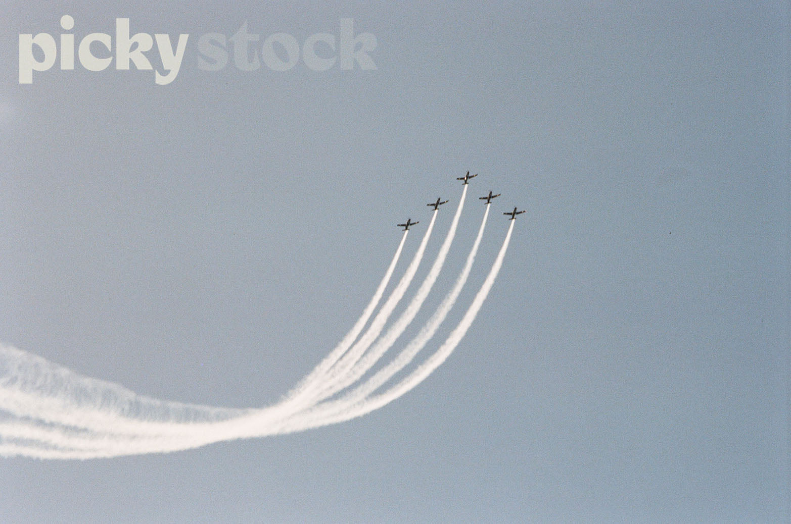 Five planes flying in the in the sky in formation, with a trail of smoke. Airshow display in Tauranga. Sky is blue and cloudless. 