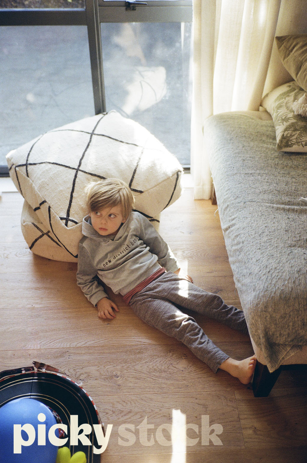 Small boy in home clothes leaning against two bean bags, looking off camera. He is sitting on a wooden floor, in a lounge / living room. Grey couch to the right with a stack of cushions. Black sliding doors and windows are behind him. 